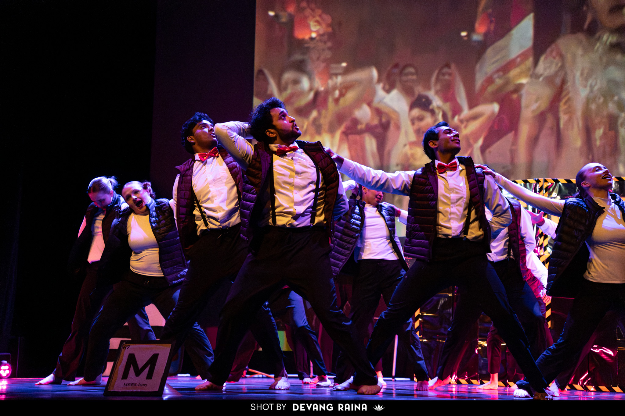 Group of dancers performing on stage with colorful background, wearing formal attire, in a synchronized dance pose.
