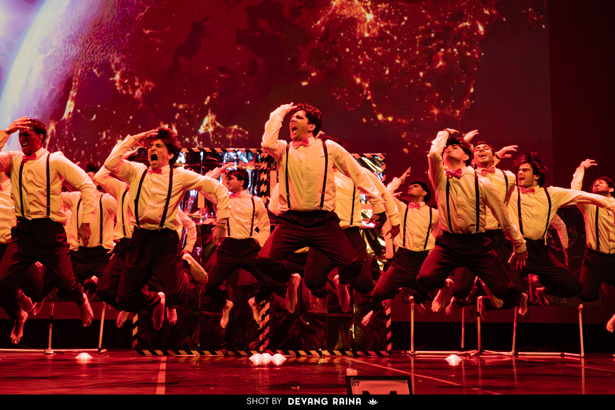 Group of performers in yellow shirts and black pants dancing on stage with a fiery background.