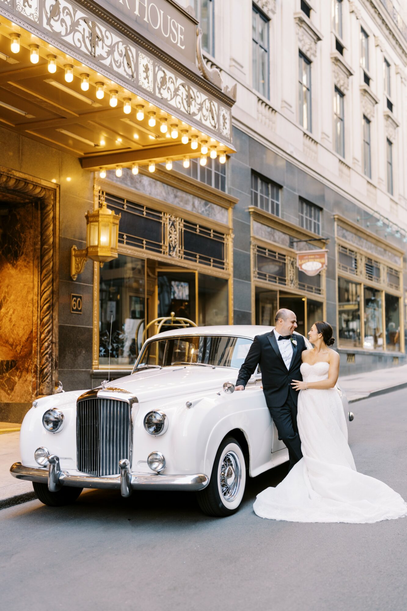 Couple in wedding attire standing beside a vintage white car on city street, building with large windows behind them.