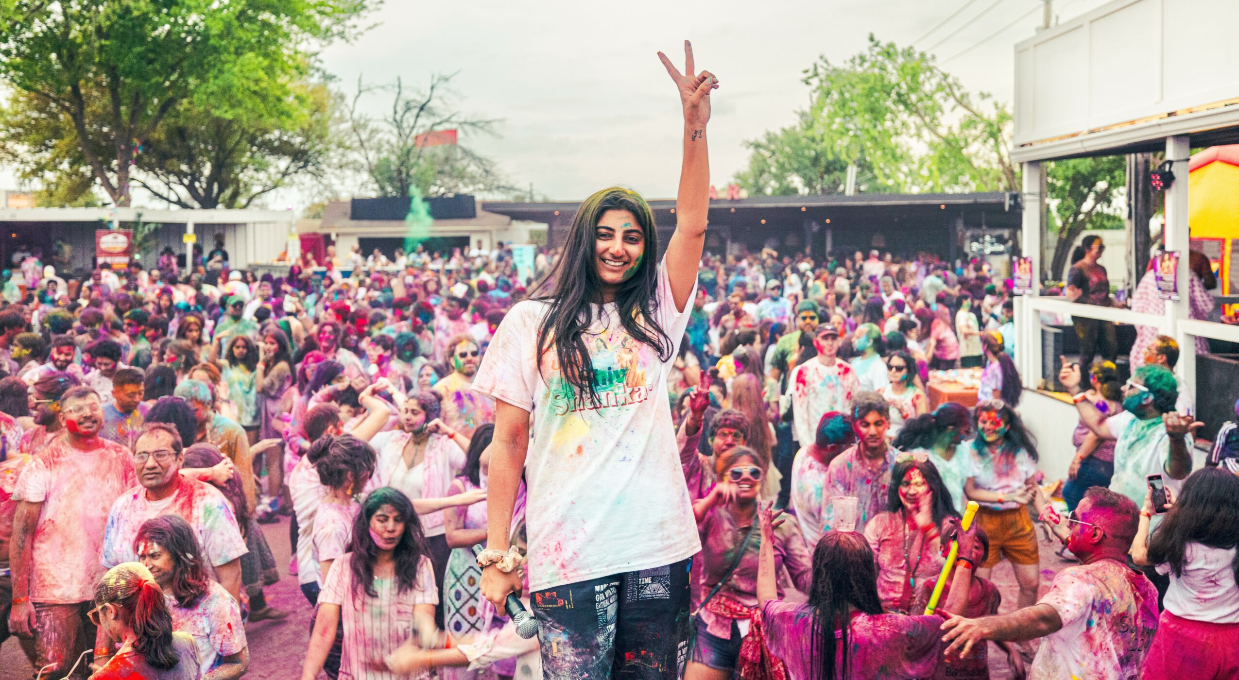 Woman standing among a large crowd of children outdoors, raising her hand in a peace sign, colorful scene.