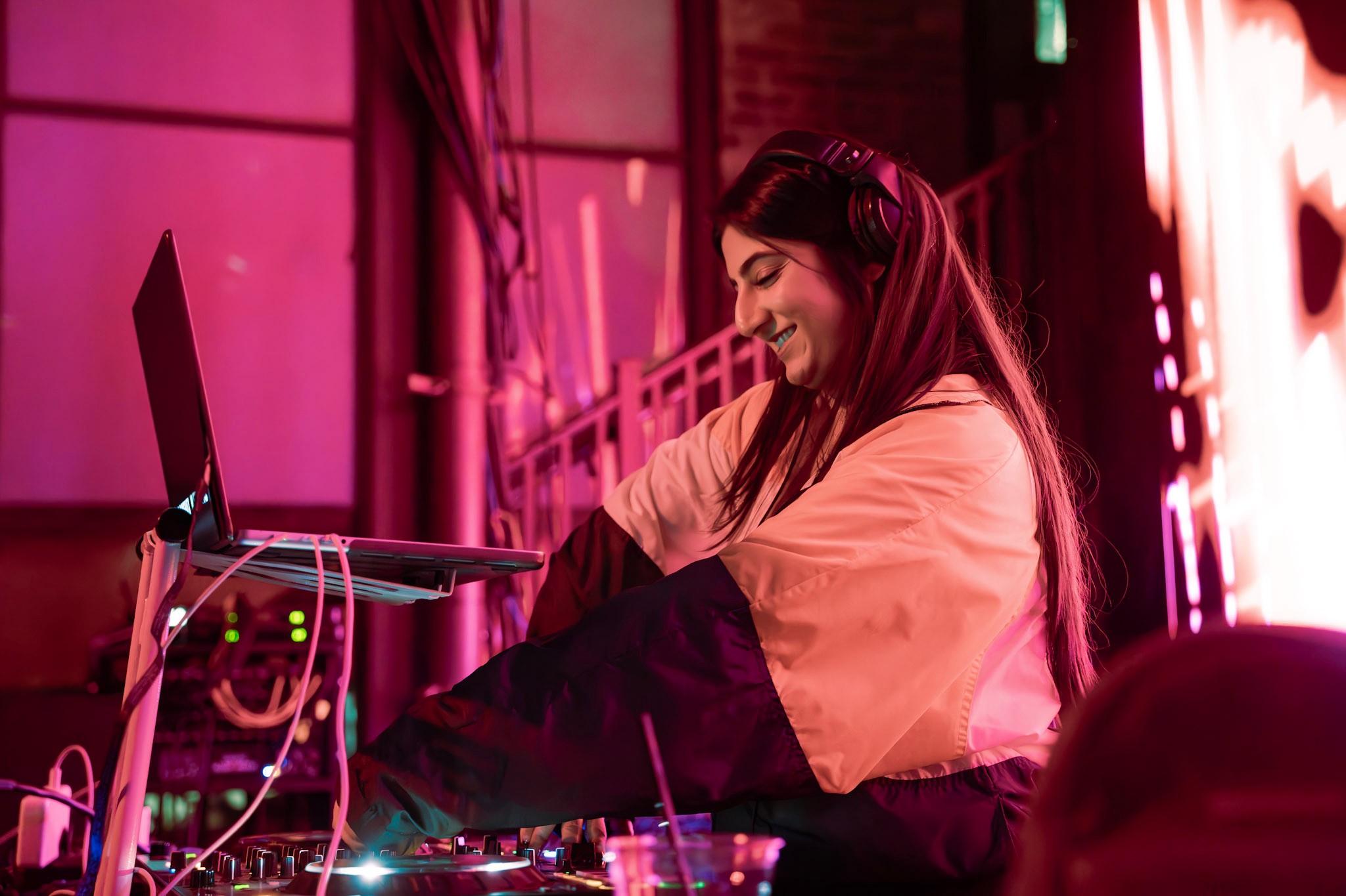 Young woman smiling at DJ equipment in a colorful, illuminated room with pink and purple lighting.