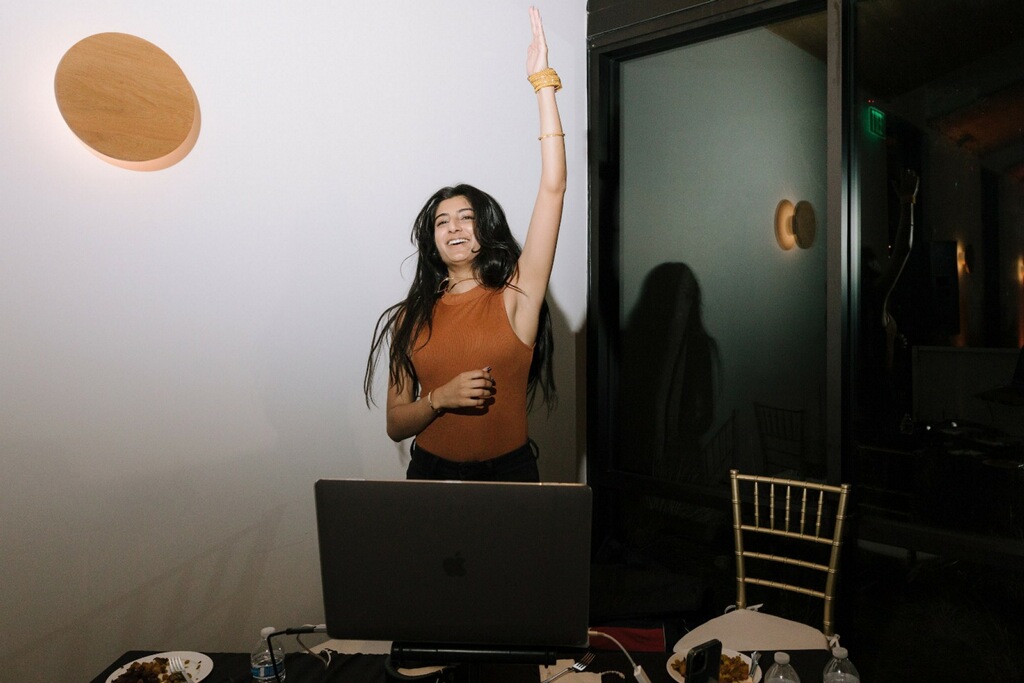 Woman with long dark hair raising her arm, standing near a laptop on a table, in a room with white wall and black door.