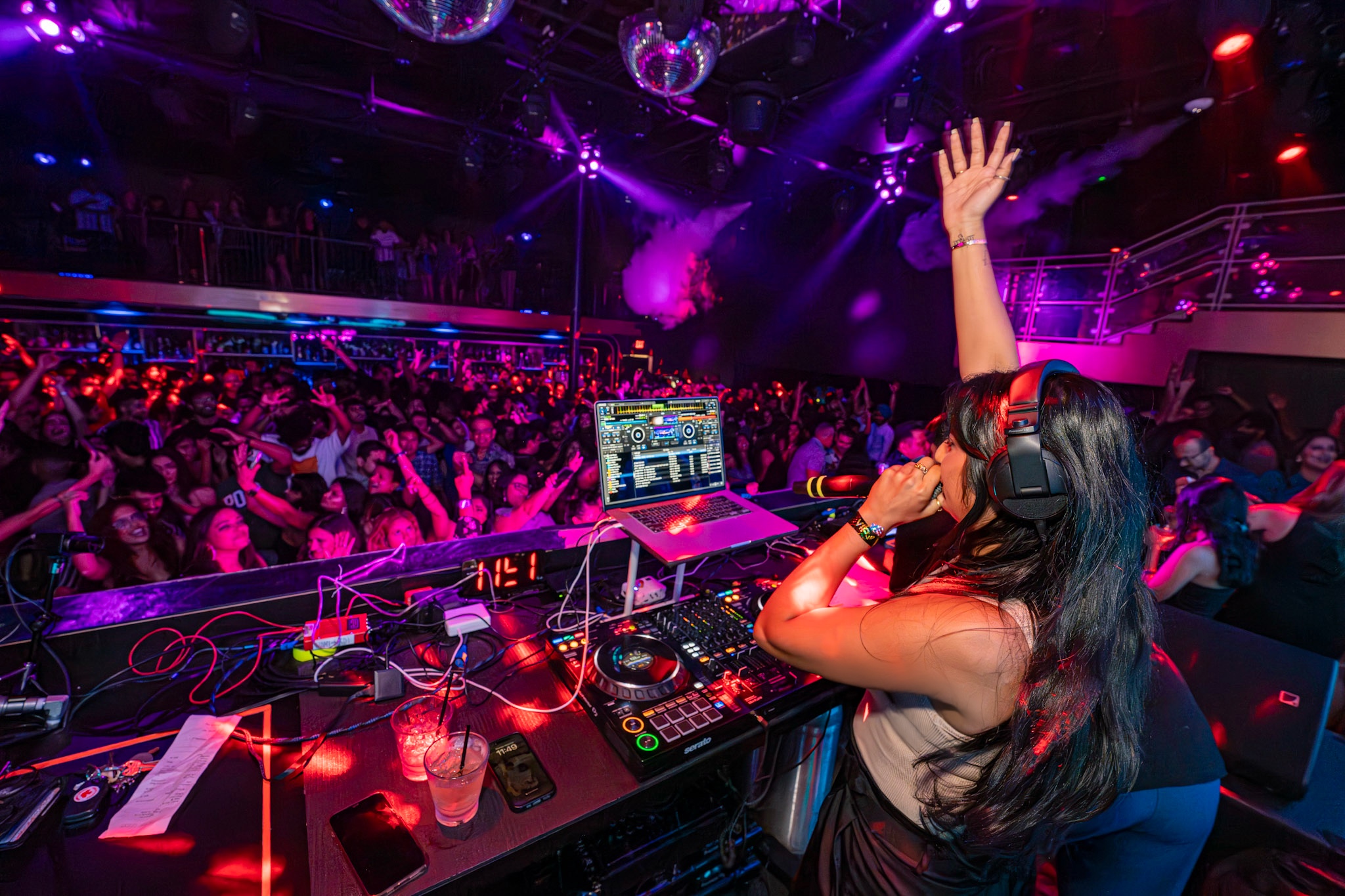 DJ with long dark hair and headphones raises hand in a crowded nightclub with colorful lights.