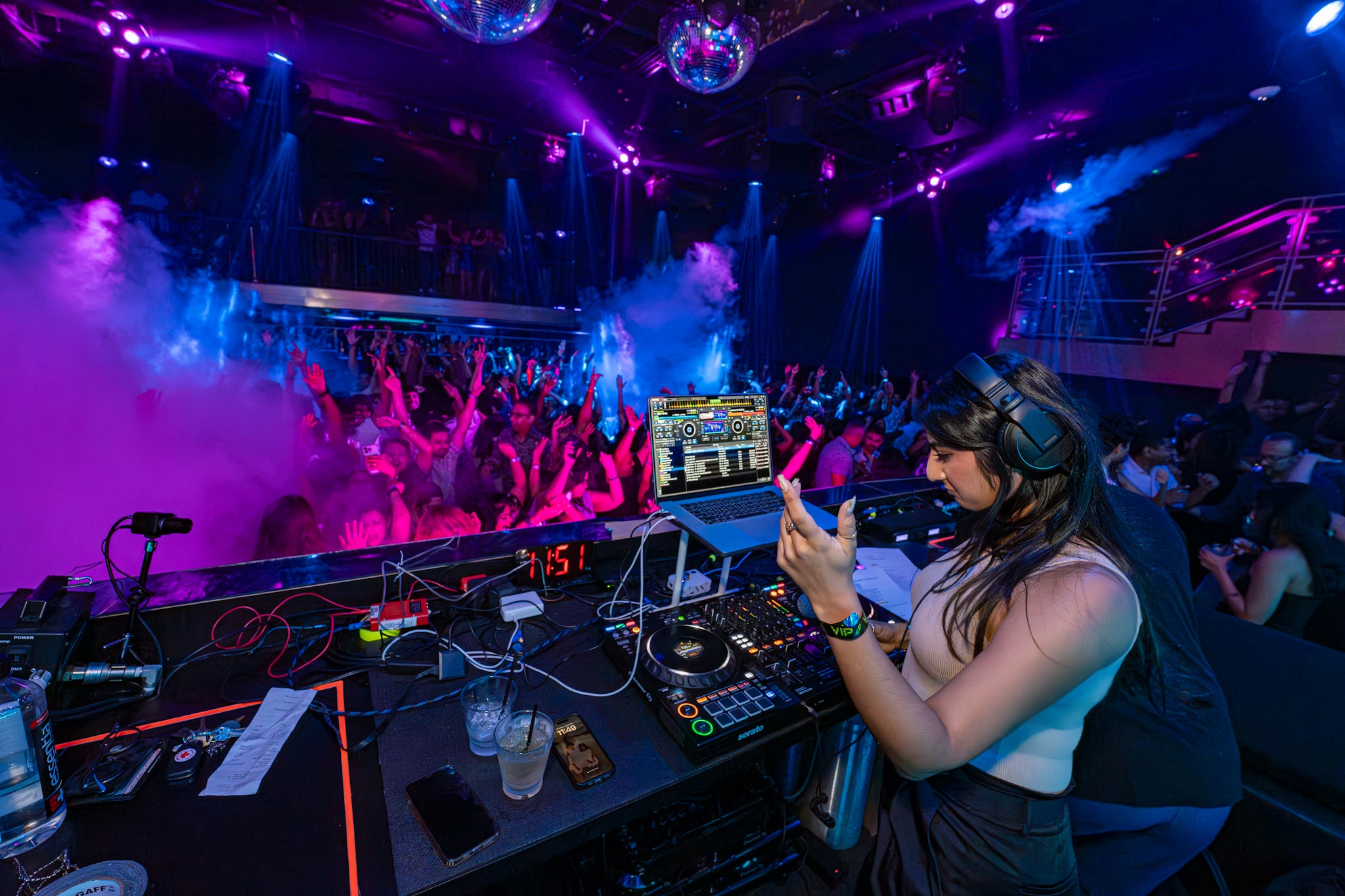 DJ woman wearing headphones using a laptop and mixer at a nightclub with colorful lights and a crowd dancing.