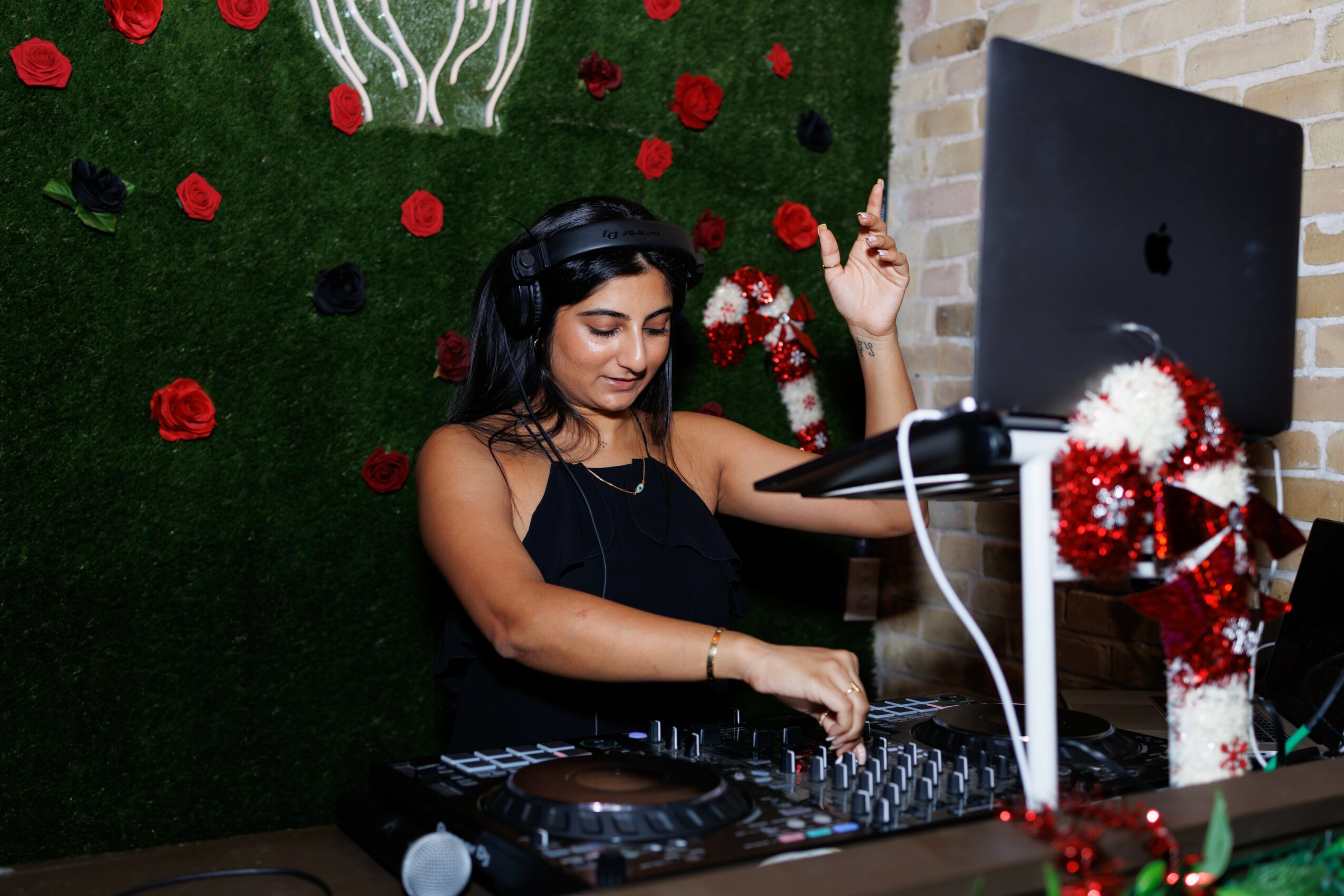 Woman DJ wearing headphones, mixing music at a holiday-themed setup with Christmas decorations, computer, and festive background.