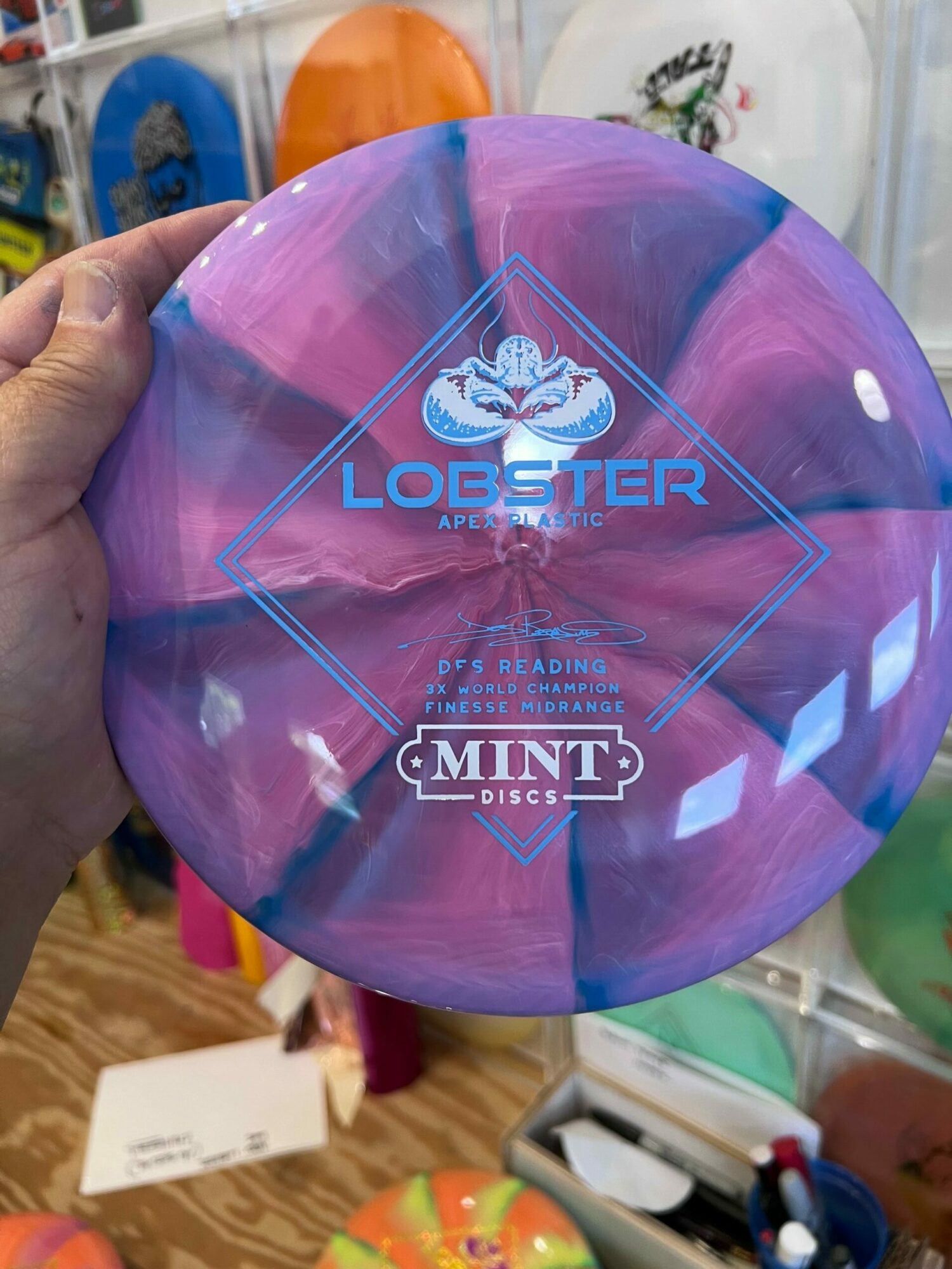 Purple and pink marbled disc with logos and text, held by a hand, on a cluttered table.