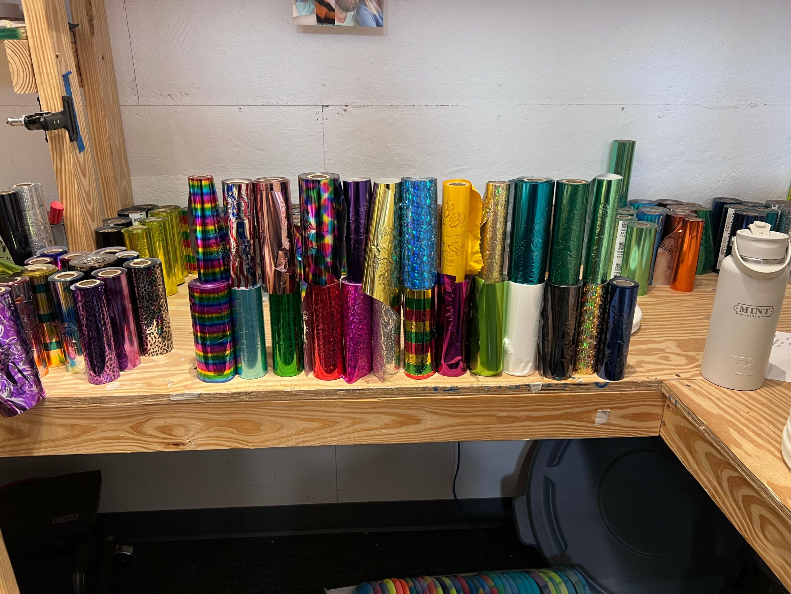 Colorful rolls of wrapping paper arranged on a wooden table, with a white wall and a water bottle in the background.