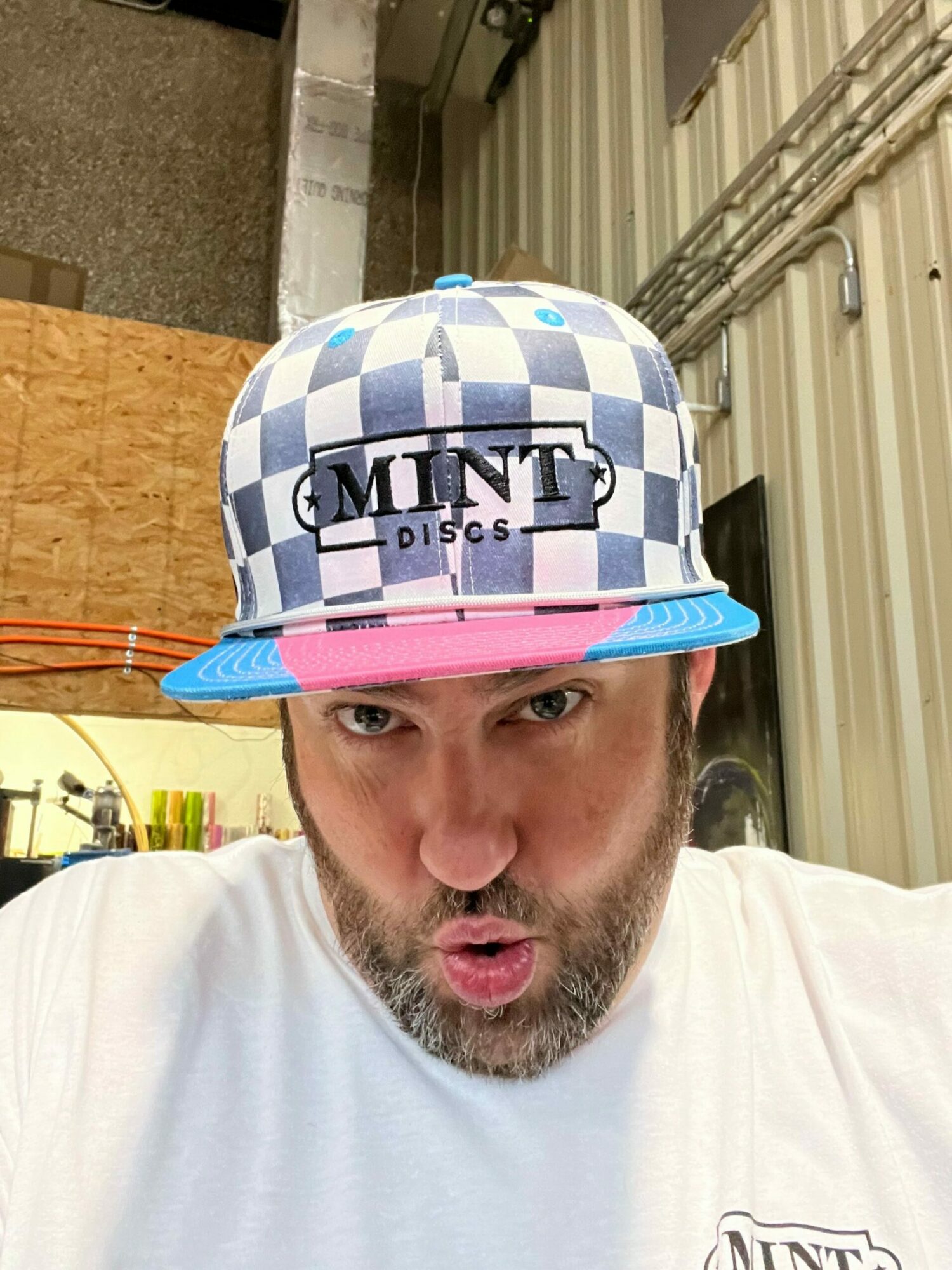 Man with beard wearing a checkered cap and white shirt, looking at the camera indoors.