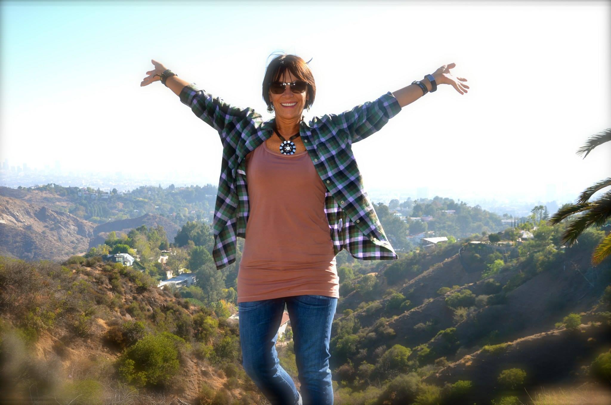 Person standing outdoors with arms outstretched, hills and sky in background, smiling, wearing sunglasses, plaid shirt, and jeans.