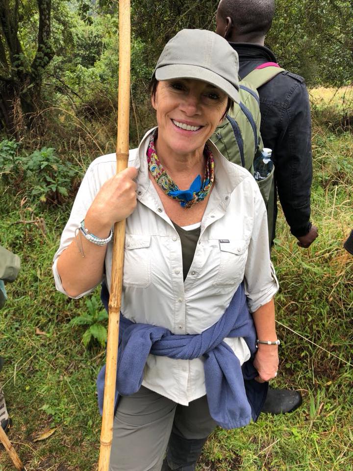Smiling woman outdoors holding a wooden stick, wearing a hat, white shirt, and colorful necklace, with a backpack and another person in background.