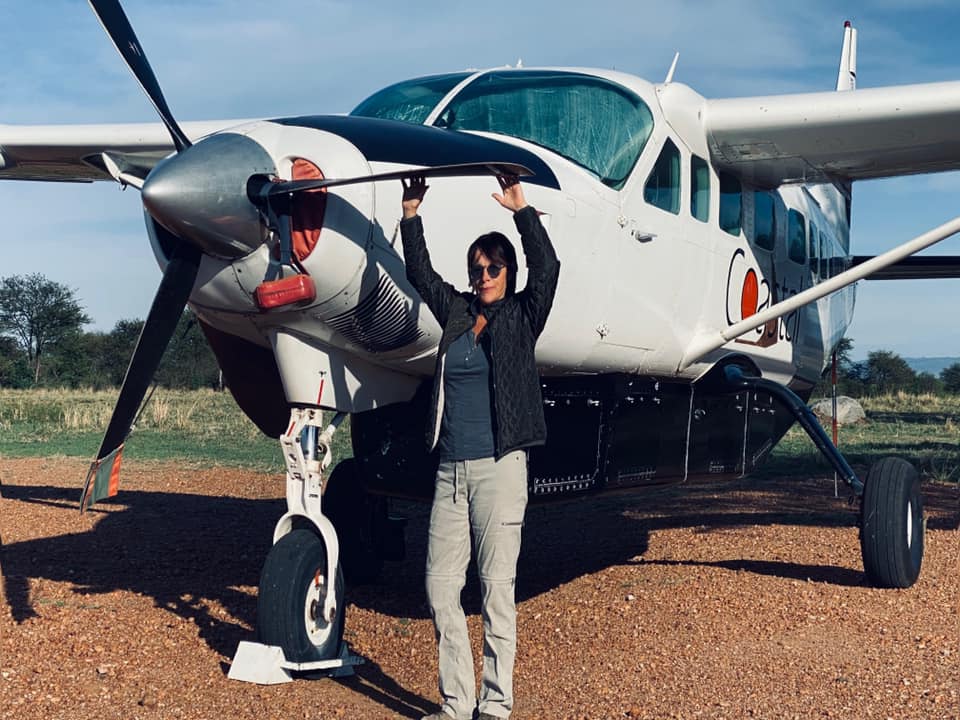 Person standing in front of a small white airplane with a propeller, holding onto the plane's wing, outdoors on a clear day.