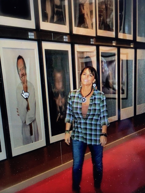 Woman standing in front of framed photographs on a wall, smiling, wearing a plaid shirt and jeans.