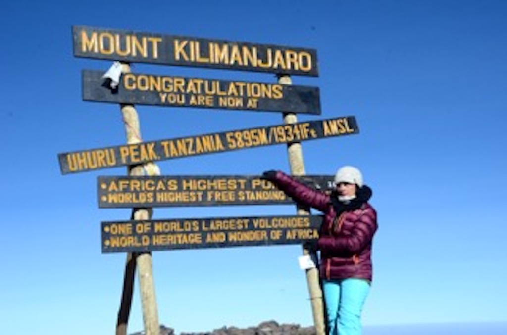 Person in purple jacket and white hat pointing at a signpost with multiple signs about Mount Kilimanjaro and Tanzania, against a blue sky.
