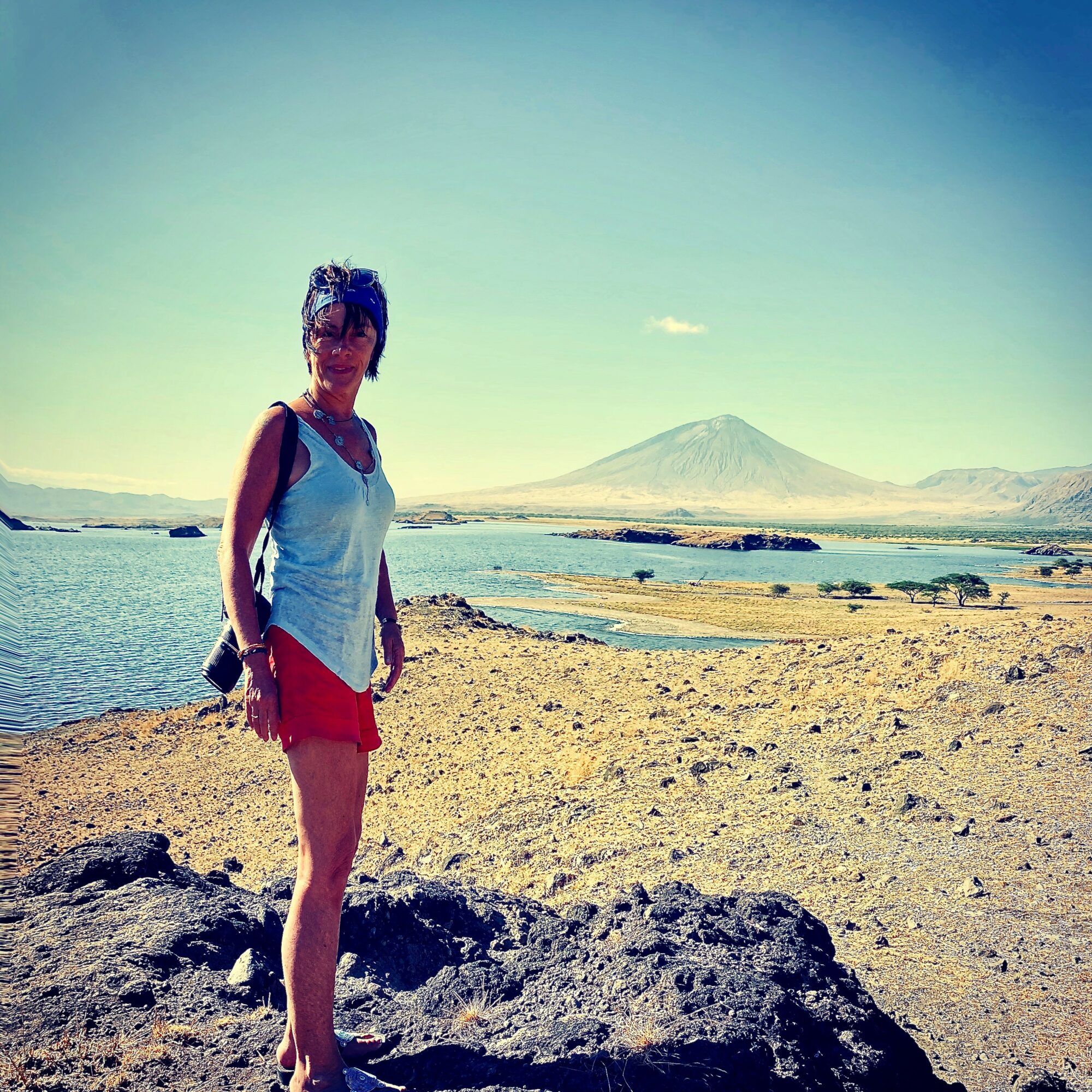 Woman standing on rocky shore with water and mountains in background, clear sky, wearing sunglasses, tank top, and shorts.