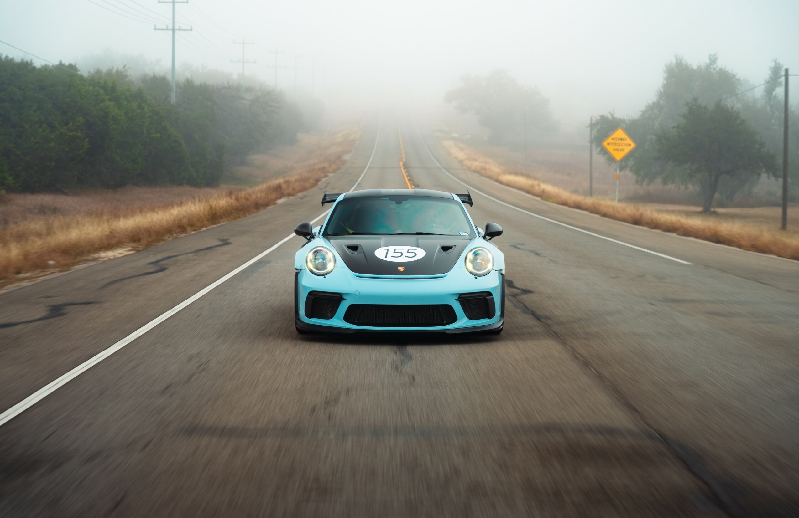 Blue sports car on a straight road with foggy background and trees on sides.