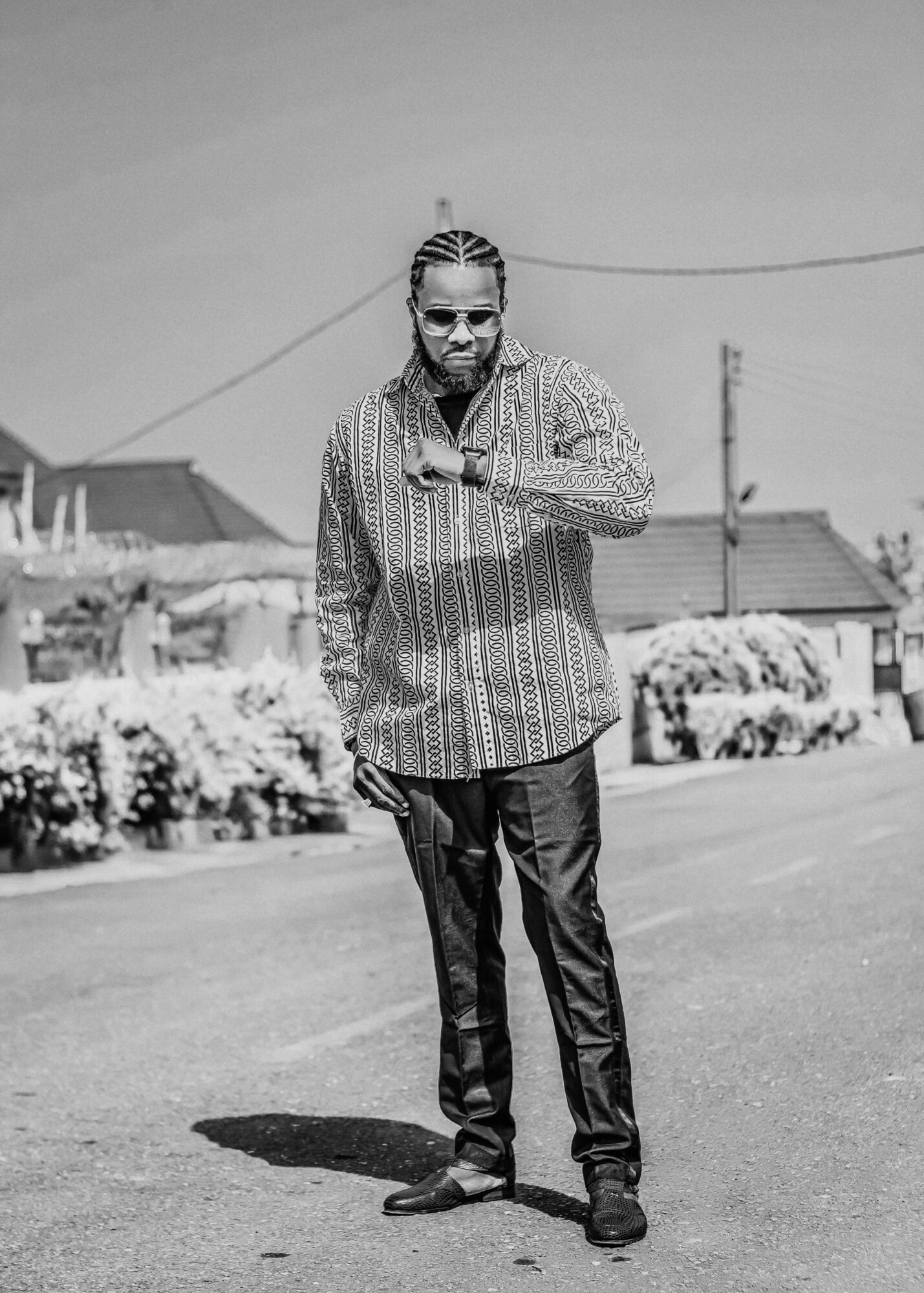 Man with braided hair and patterned shirt walking on street with houses and bushes in background
