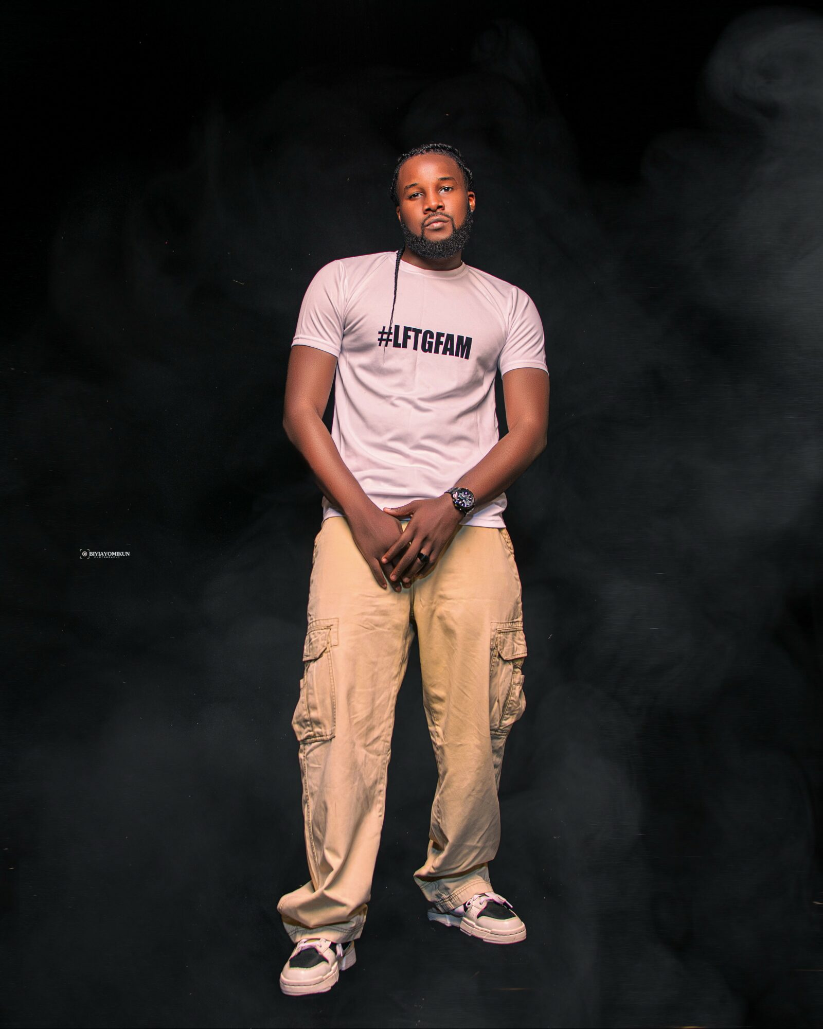 Man standing with hands clasped, wearing a light pink T-shirt with hashtag, beige cargo pants, and sneakers, against a dark background.