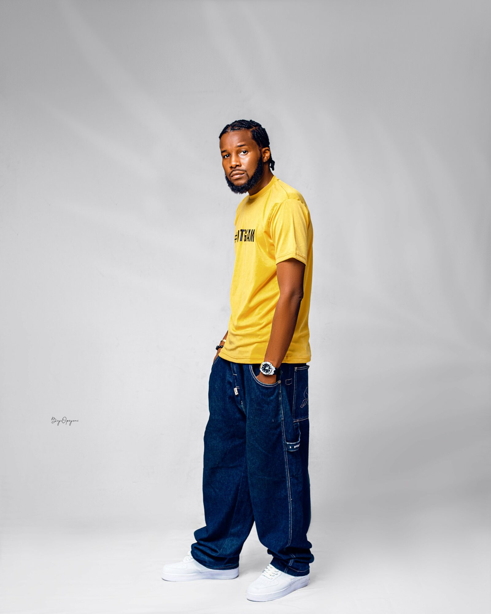 Man with braided hair and beard wearing a yellow T-shirt, baggy jeans, and white sneakers, standing against a plain background.