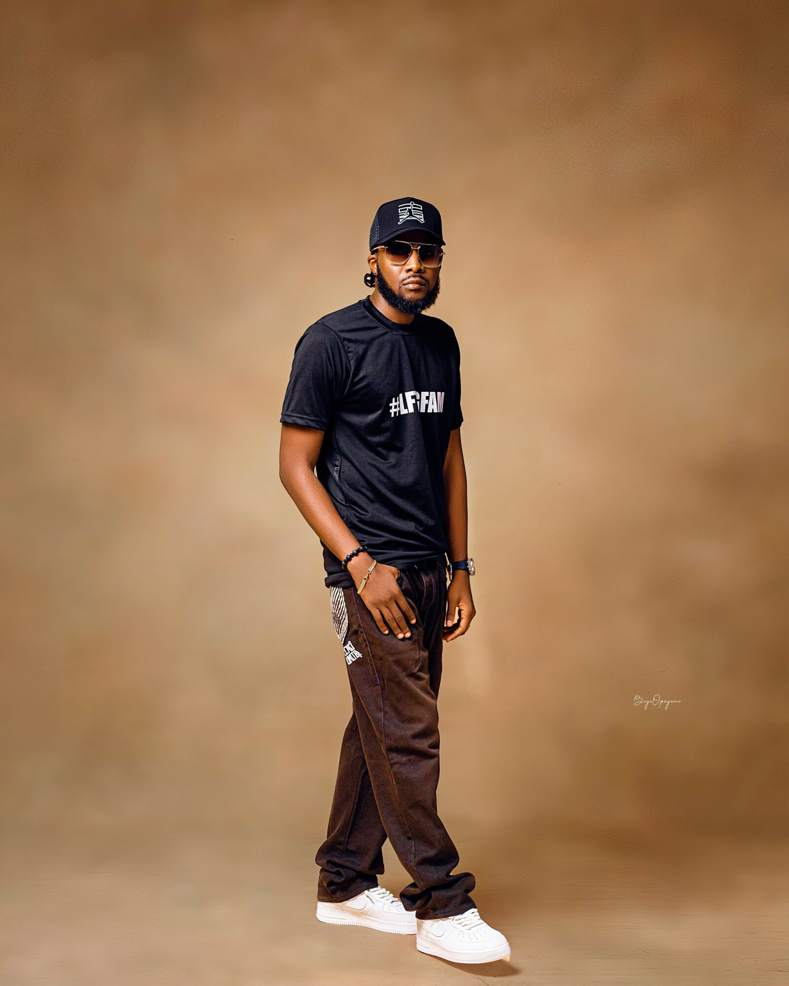 Man standing against a plain brown background, wearing a black cap, black t-shirt, brown pants, and white shoes.