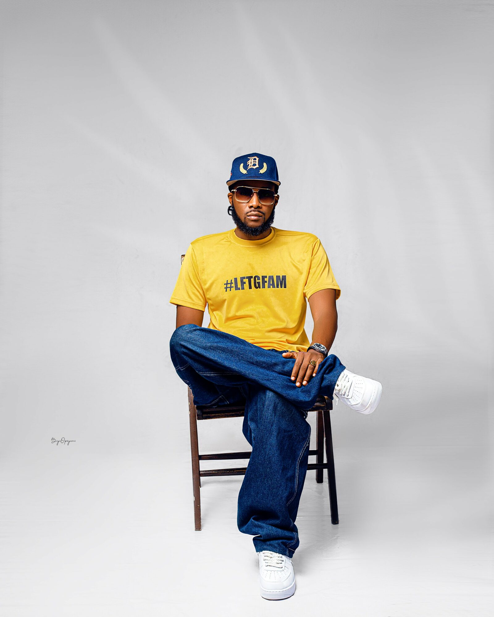 Man sitting on a chair wearing a yellow T-shirt, blue jeans, white sneakers, sunglasses, and a blue cap, against a plain background.