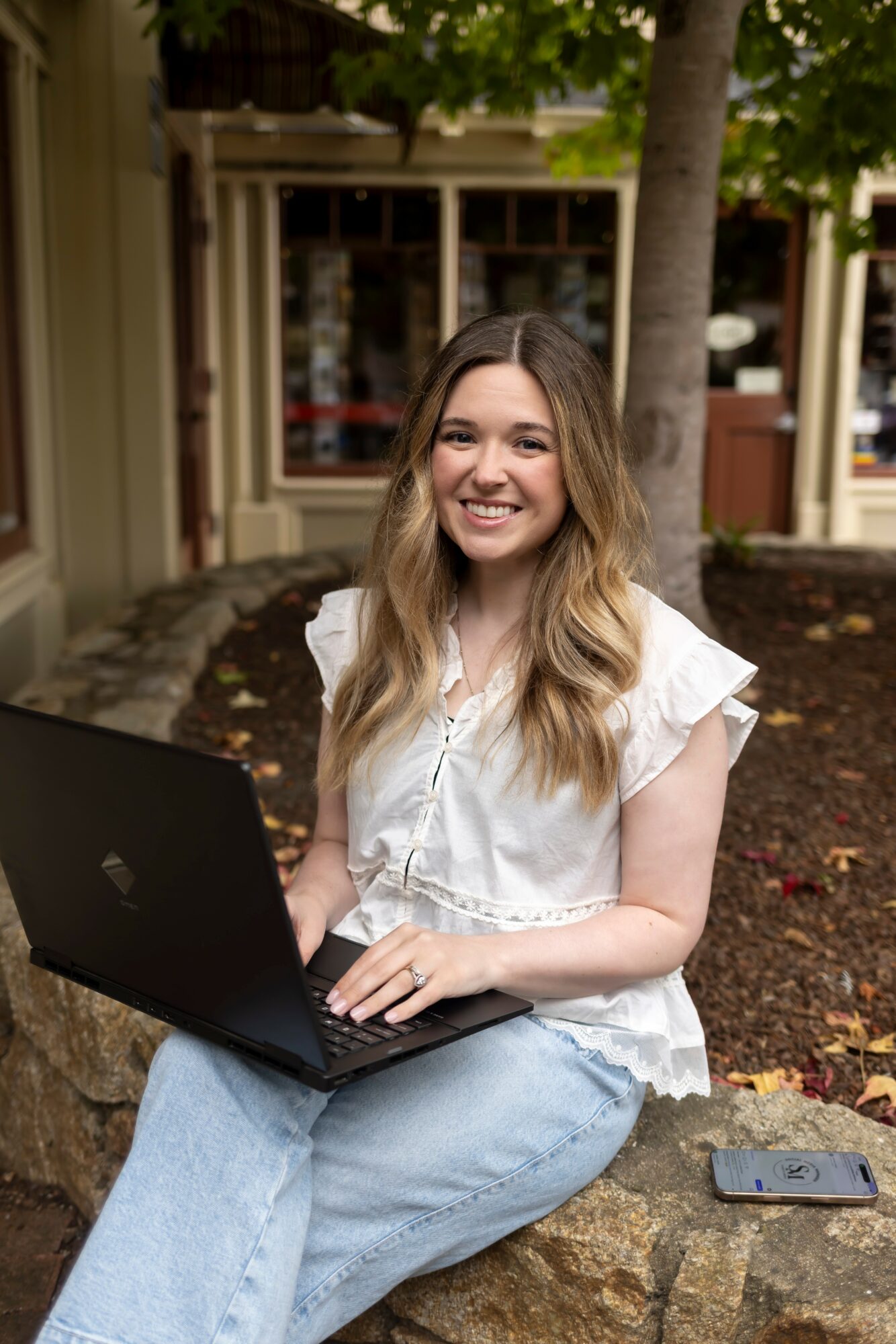Young woman with long wavy hair sitting outdoors with a laptop, smiling, in front of a building and trees.