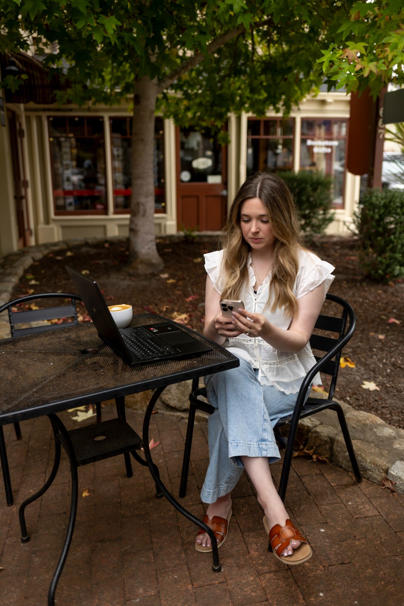 Young woman with long hair sitting outdoors at a table, using her phone, with a laptop nearby, trees and buildings in background.