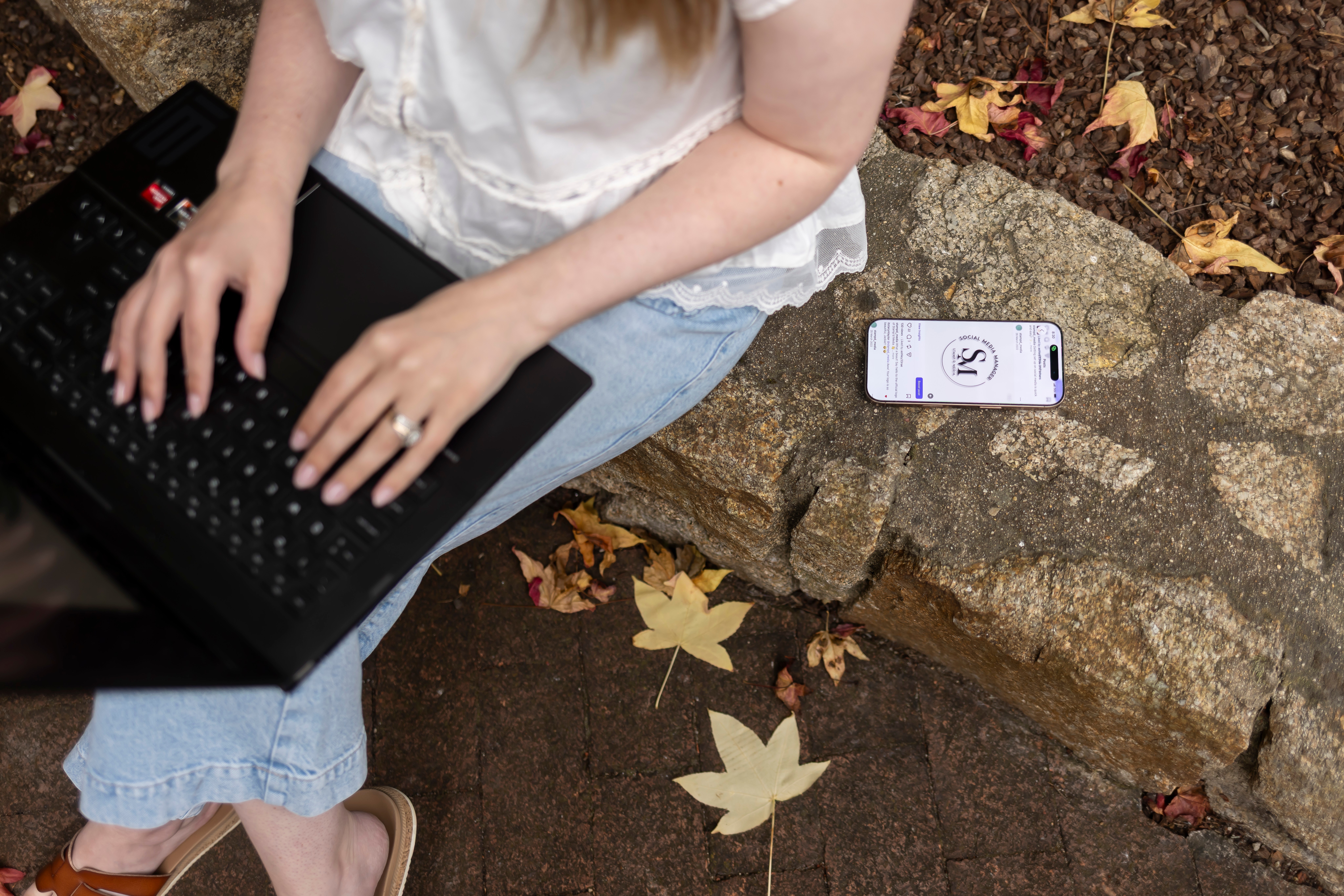 Person sitting outdoors with a laptop and smartphone on rocks, surrounded by fallen leaves.