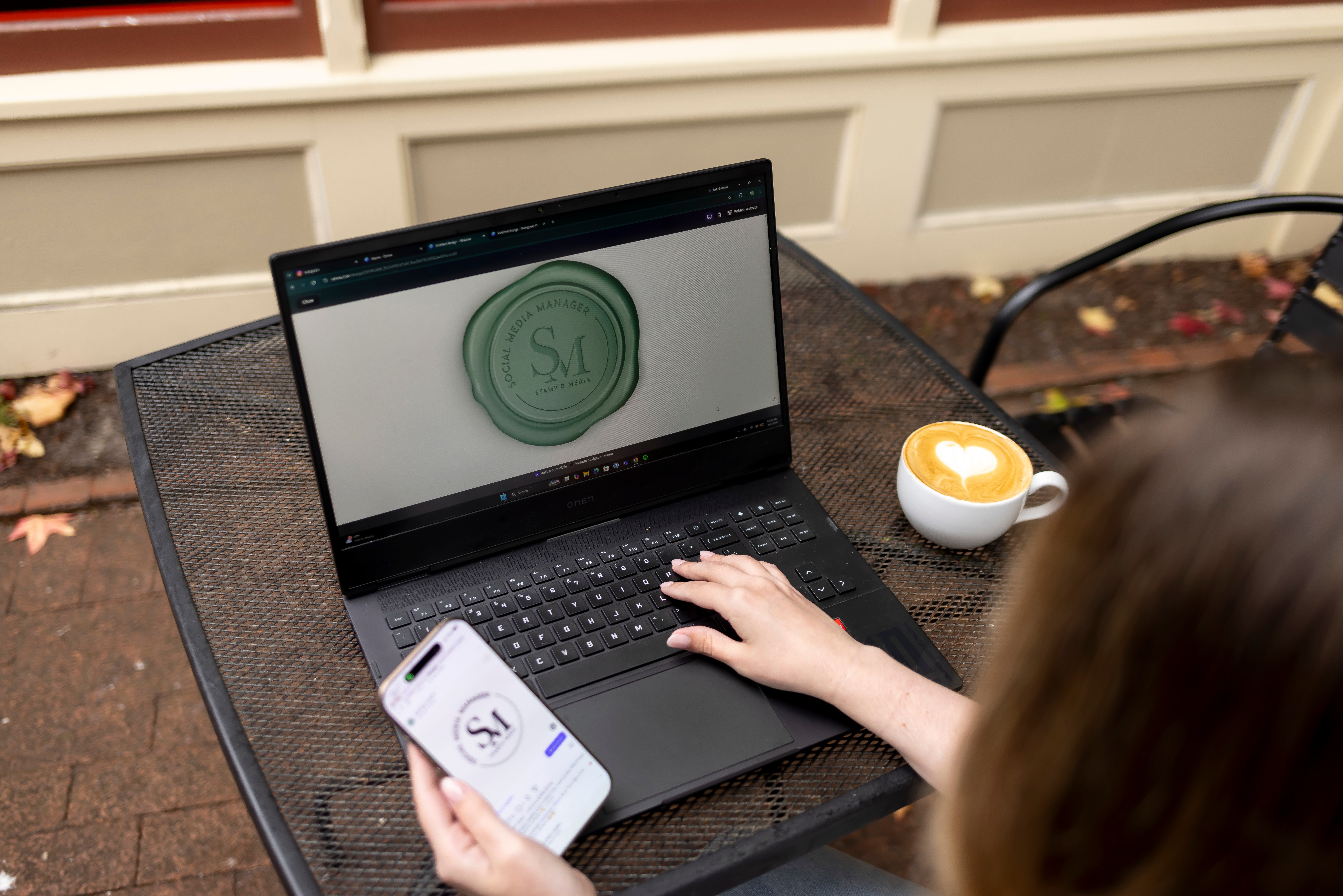 Person using a laptop outdoors, holding a smartphone, with a cup nearby on a table.