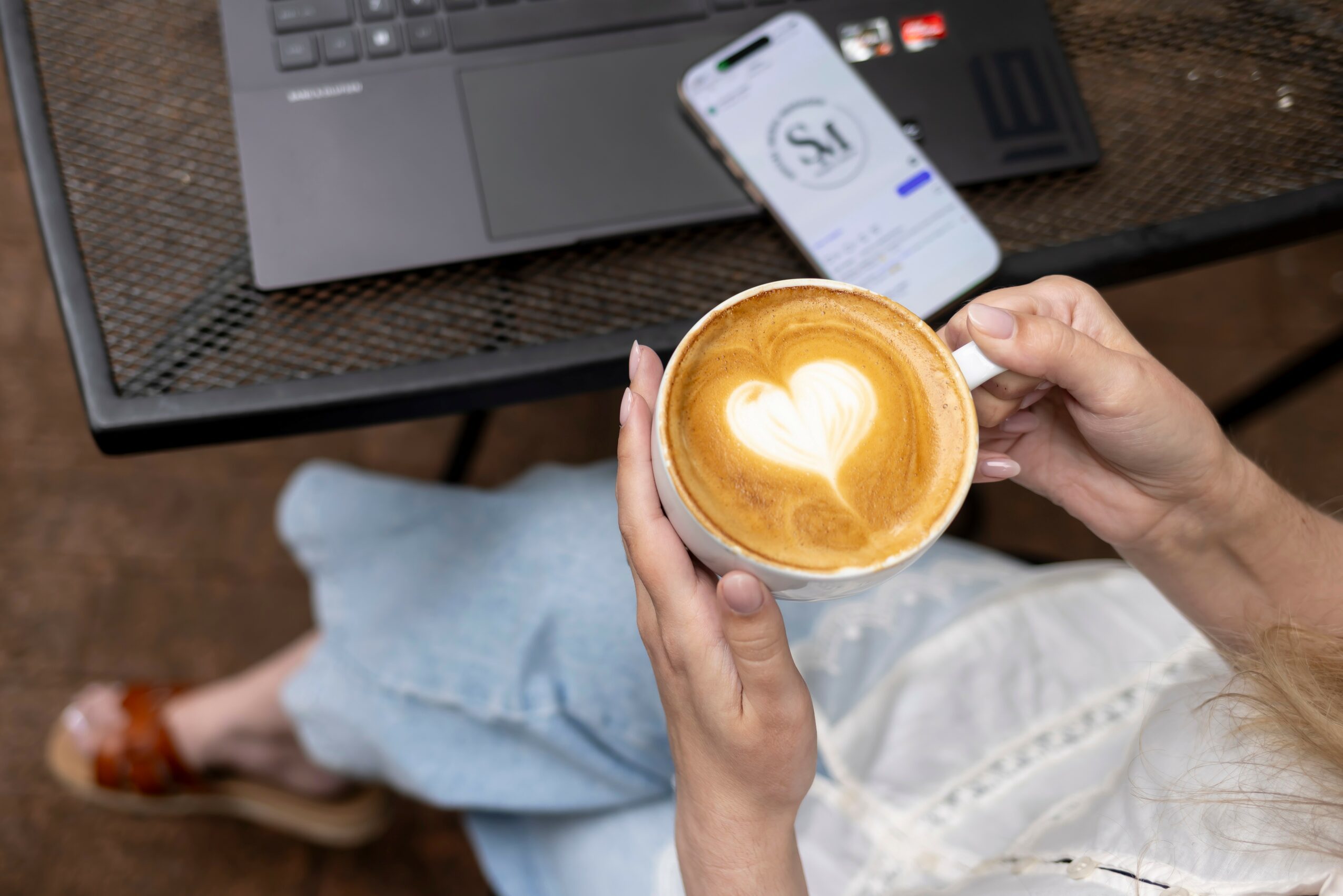 Person holding a coffee cup with a heart-shaped latte art, sitting at a table with a laptop and smartphone.