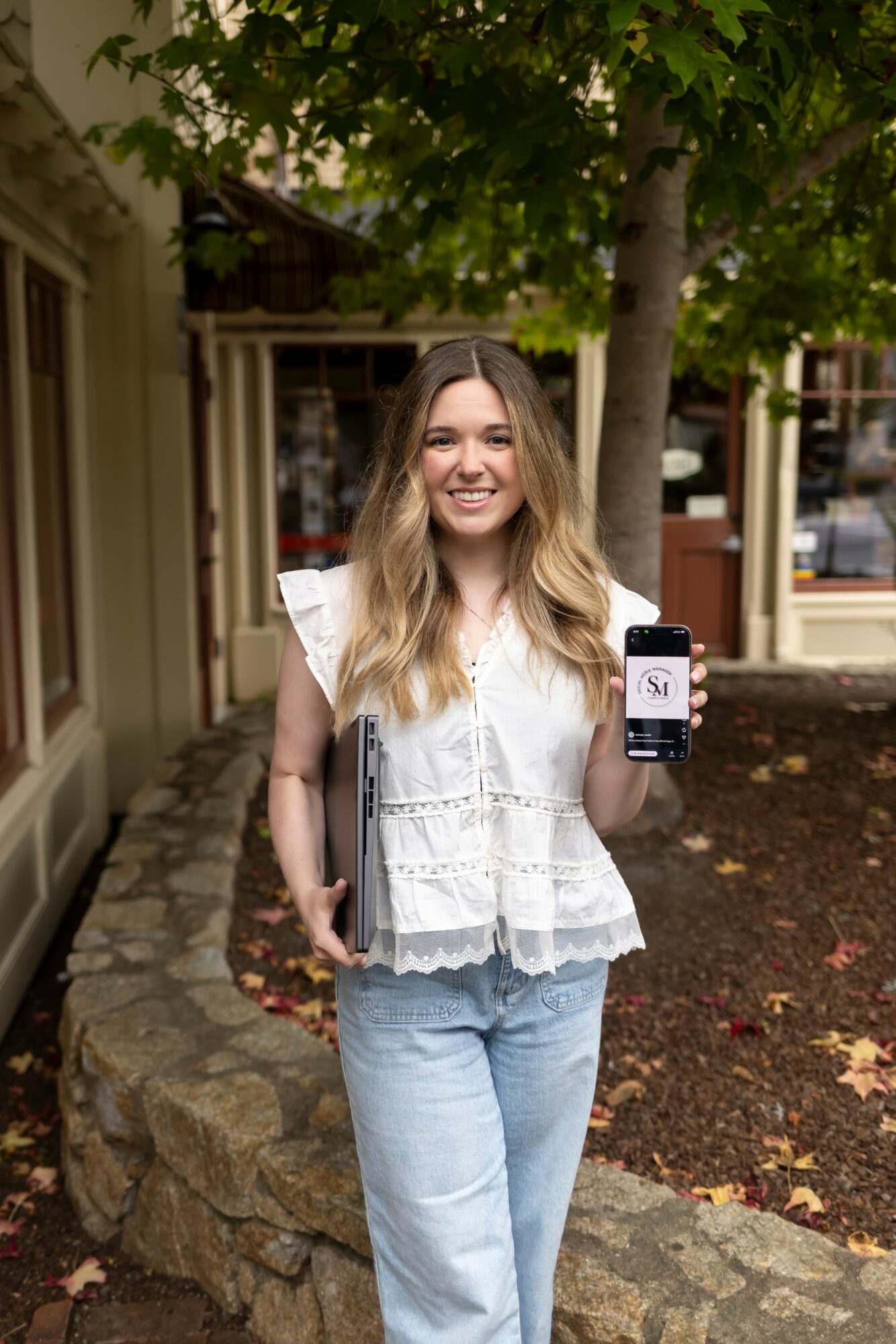 Young woman with long hair smiling outdoors, holding a smartphone showing a social media app, standing near a tree and stone wall.