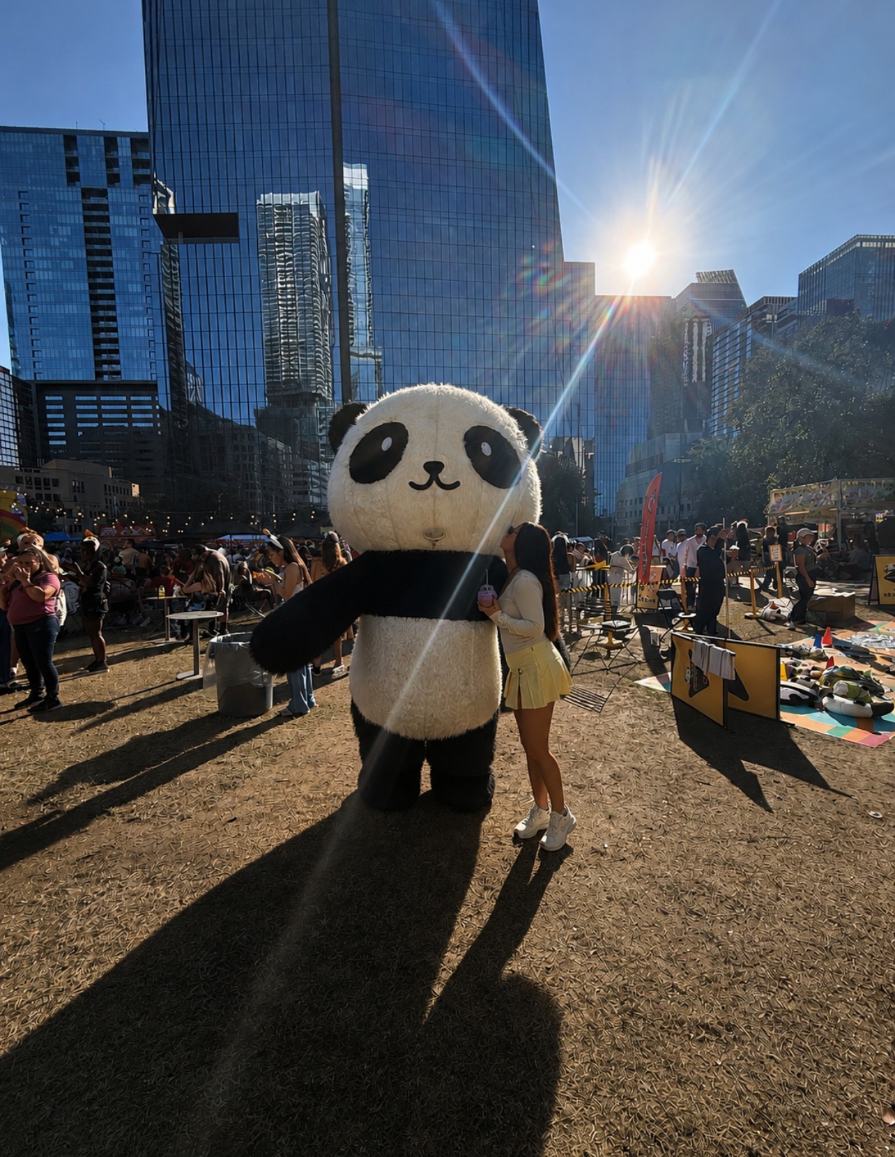 Person standing next to a large panda mascot in an outdoor urban area with tall buildings and a crowd, sunlight shining brightly.