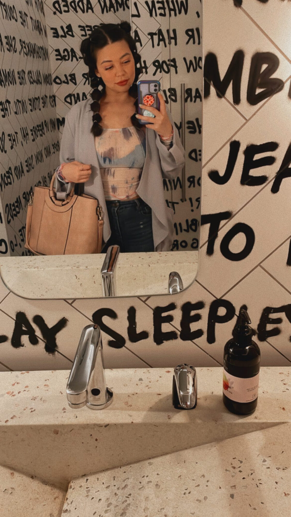 Woman taking mirror selfie in a bathroom with black and white wall art, holding a beige bag, wearing a gray jacket and jeans.