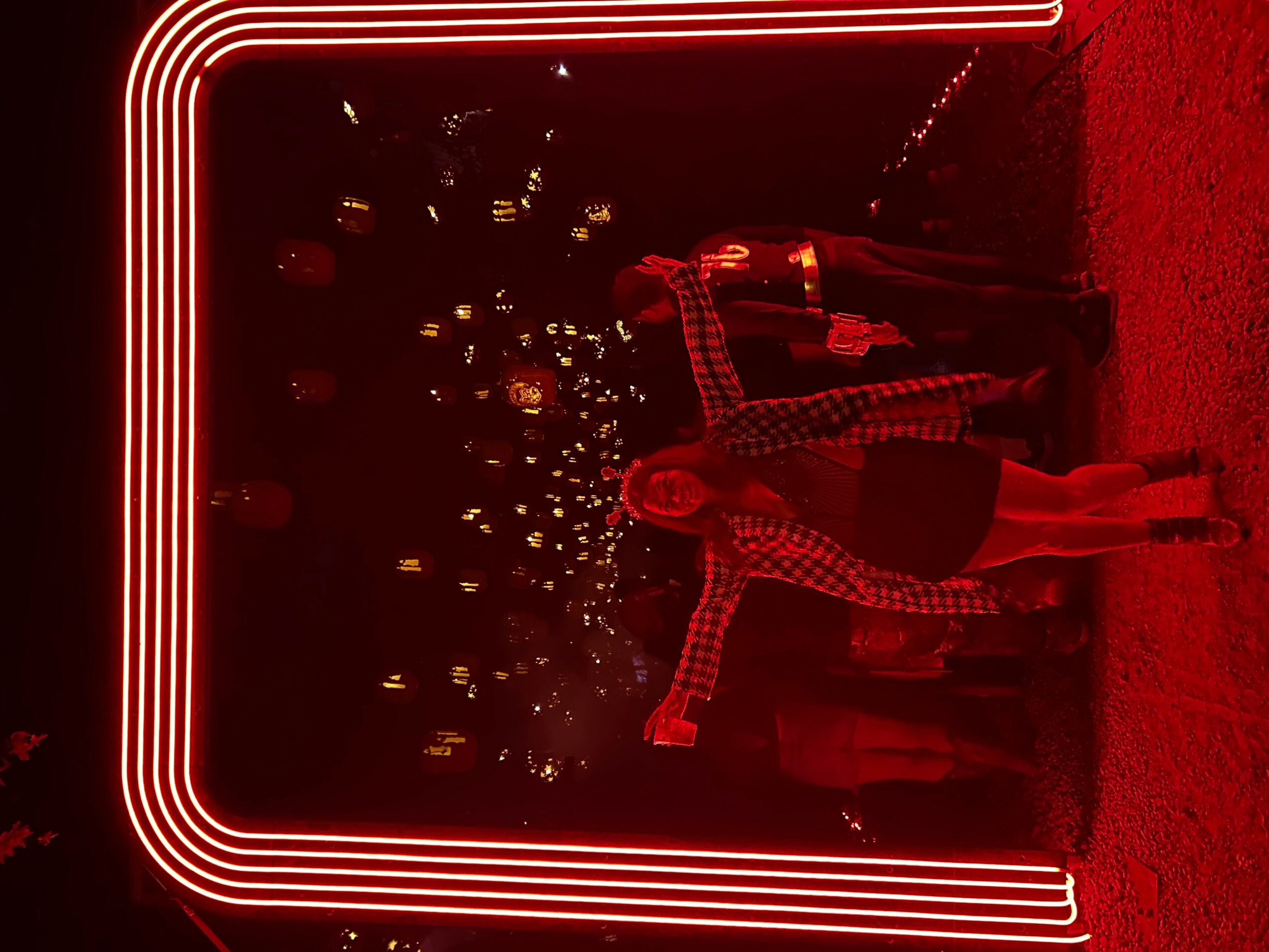Person standing inside a neon-lit frame at night, with city lights in the background, illuminated by red lights.
