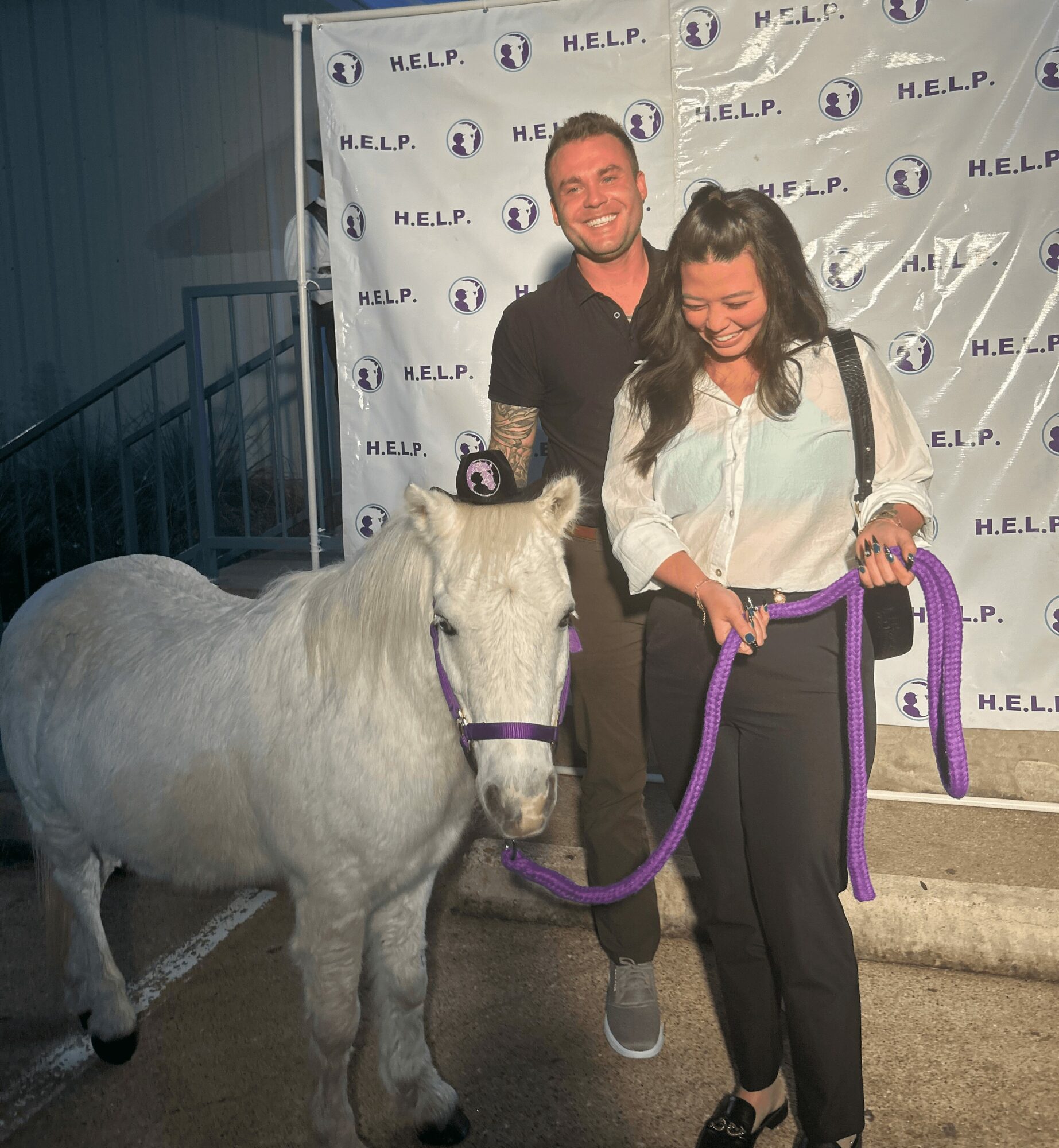 Two people smiling with a small white pony in front of a backdrop with H.E.L.P. logos, woman holding a purple lead rope.