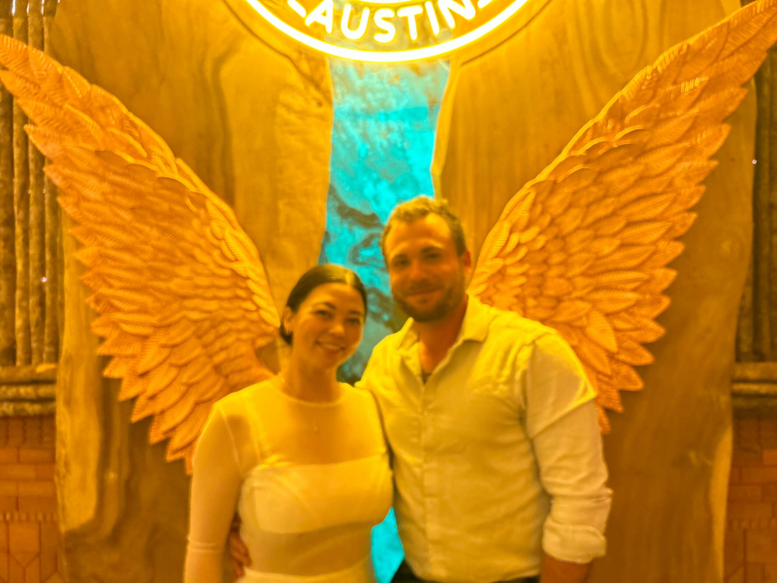 Two people stand in front of large angel wings and a sign that says 'AUSTIN'.