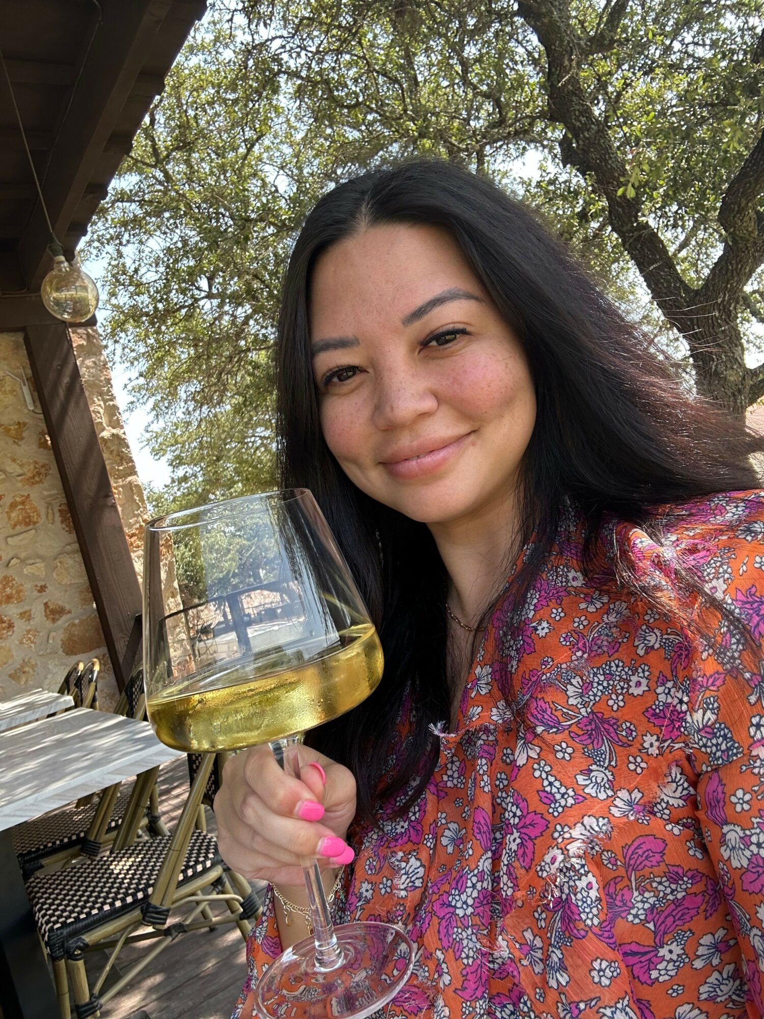 Woman with long dark hair smiling outdoors, holding a glass of beer, trees in background.