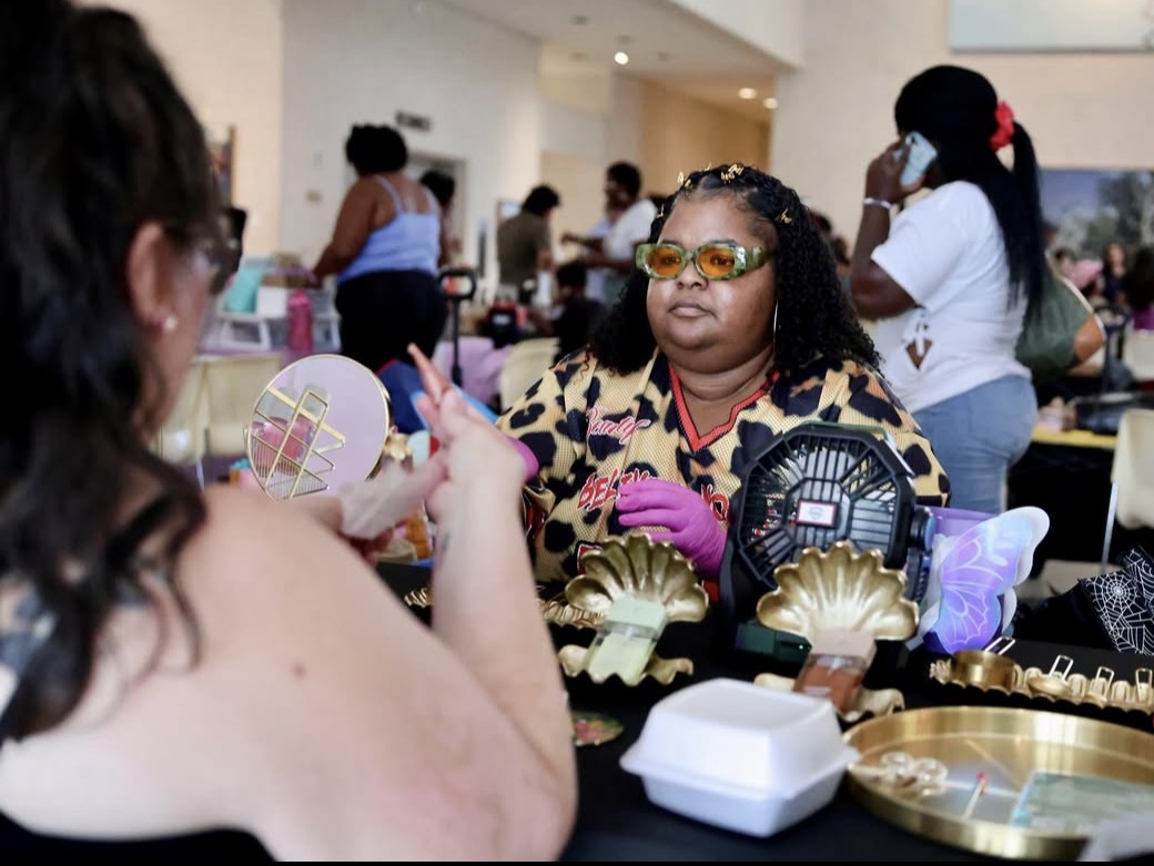Two women sit at a table with decorative items, one wearing sunglasses and gloves, in a busy indoor setting.