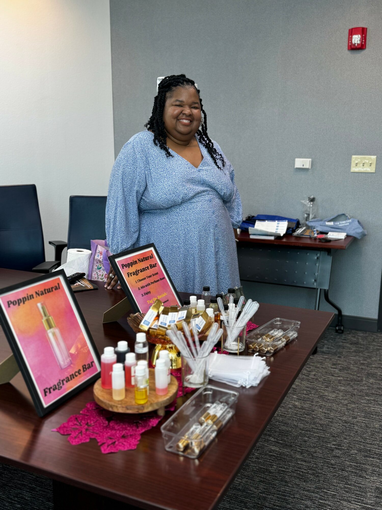 Smiling woman standing behind a table with colorful bottles and signs, in a room with gray walls and chairs.