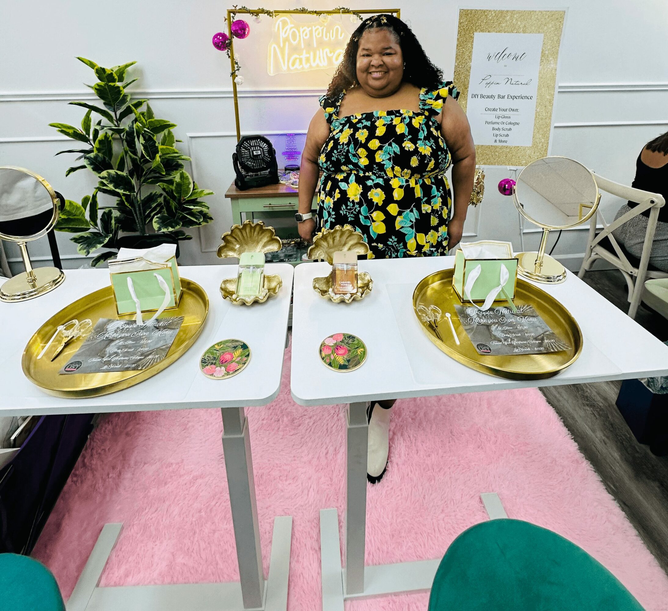Smiling woman in floral dress standing behind decorated tables with gold trays and pink accents, pink rug on floor.