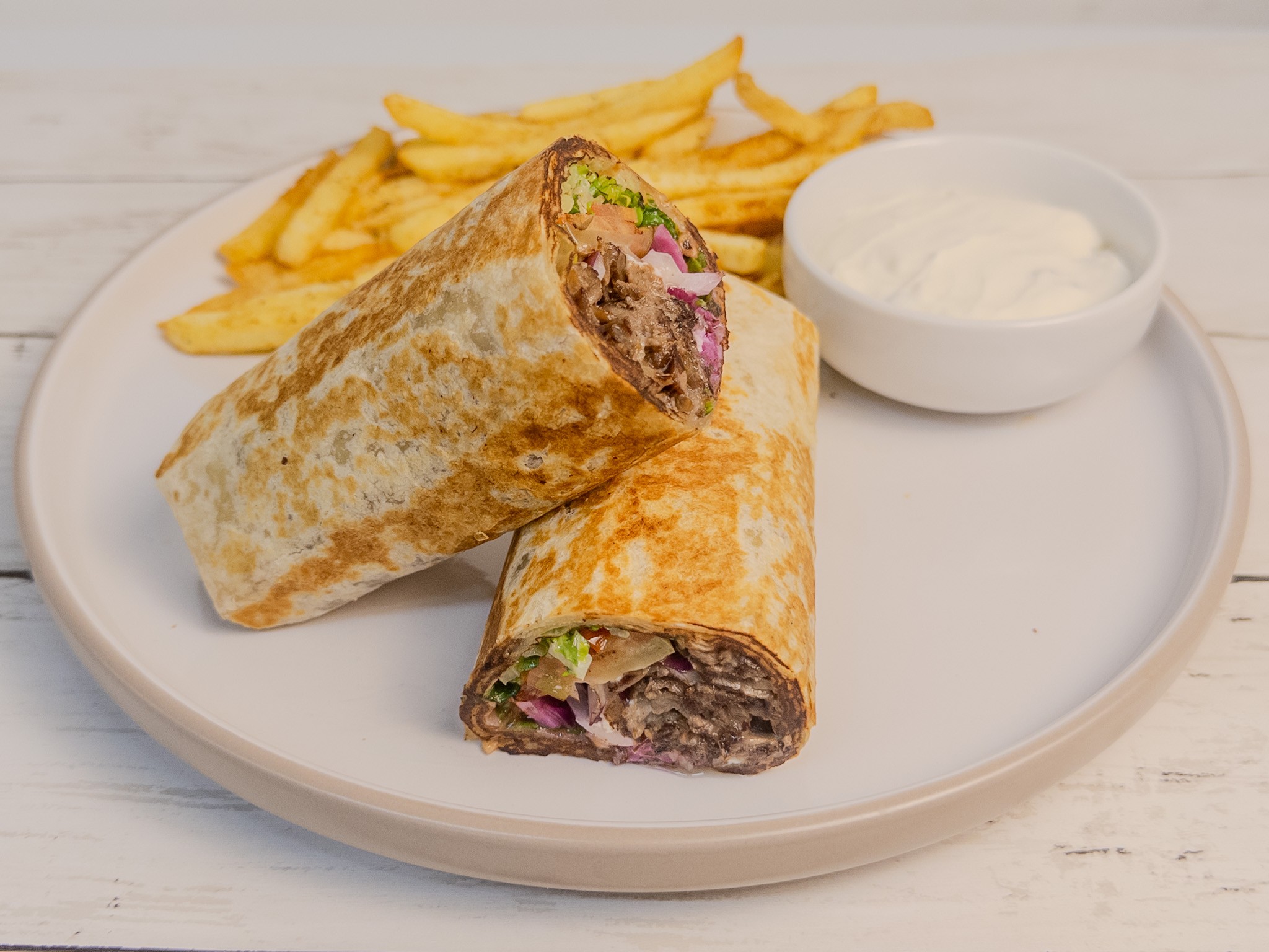 Two beef burritos on a white plate with French fries and a small bowl of sour cream, on a light wooden surface.