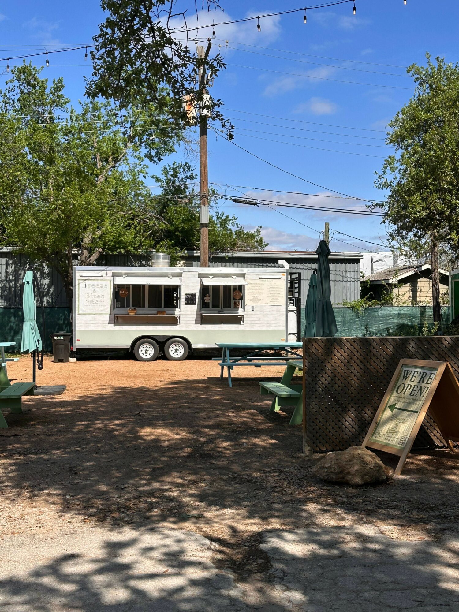 White food truck with windows and wheels parked on a street with trees and a trash bin nearby.
