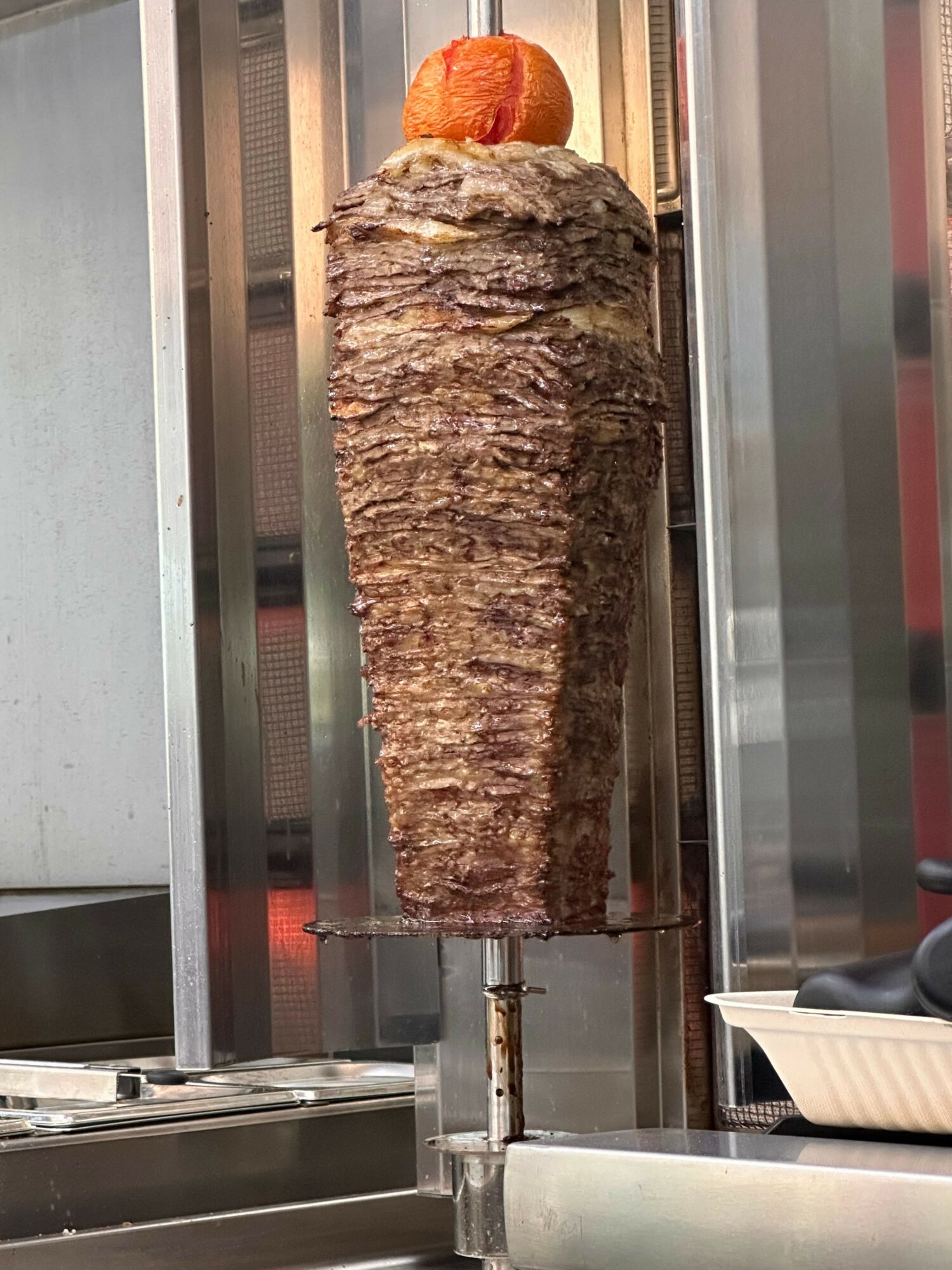 Skewered meat cooking on a vertical rotisserie with a piece of bread at the top, viewed sideways.