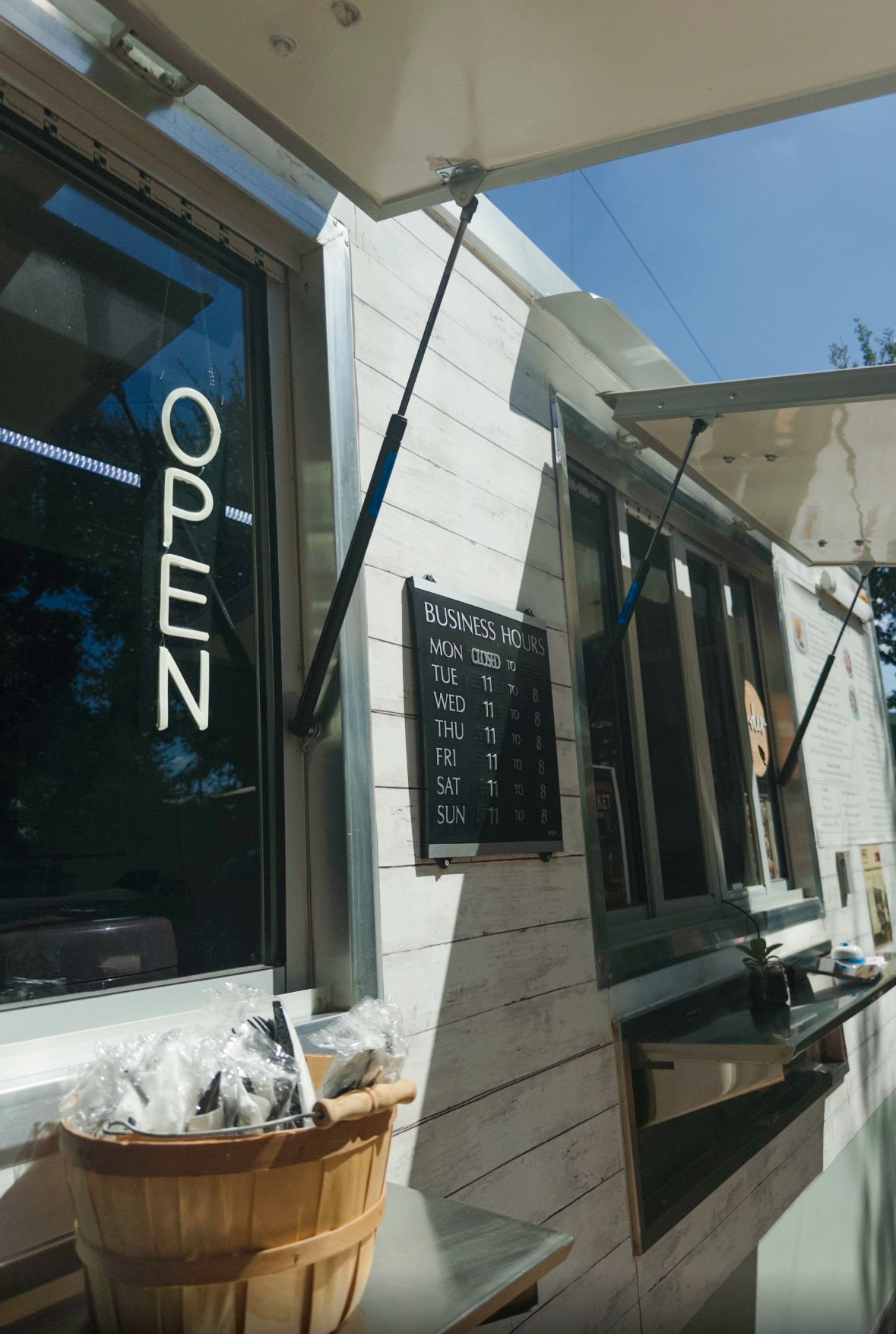 Outdoor cafe with an open sign, menu board, and umbrellas, against a building with windows and a clear blue sky.