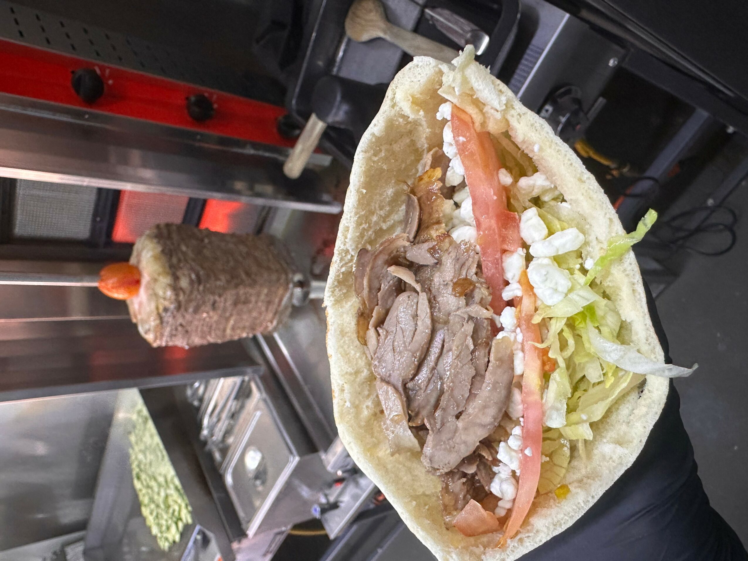 Sandwich with sliced meat, lettuce, tomato, and sauce in a bun, held in hand. Background shows a grill and a rotisserie with meat.