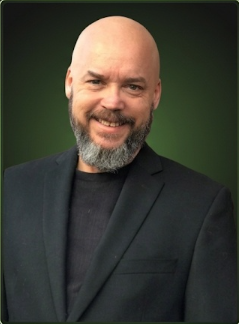Bald man with beard smiling, wearing a black blazer and shirt, against a green background.