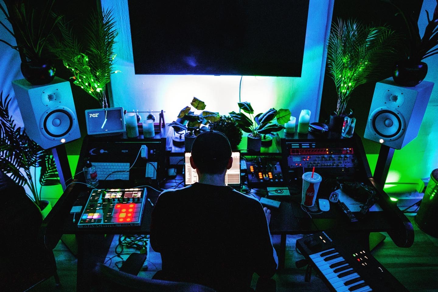 Person working at DJ or music production setup with speakers, monitors, and equipment, illuminated by colorful lighting, with plants and a large screen behind.