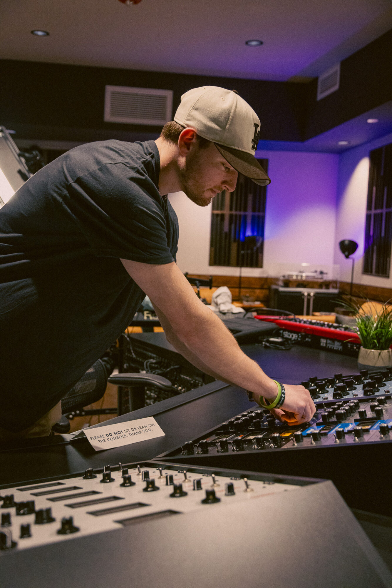 Person wearing a cap and glasses adjusts equipment at a DJ setup in a dimly lit room with purple lighting.
