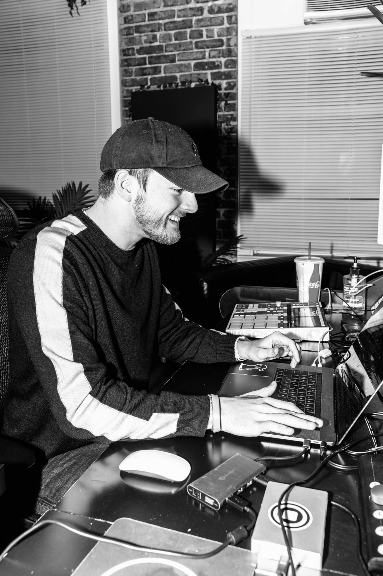 Person wearing a cap and glasses working on a laptop at a desk with electronic devices and drinks.