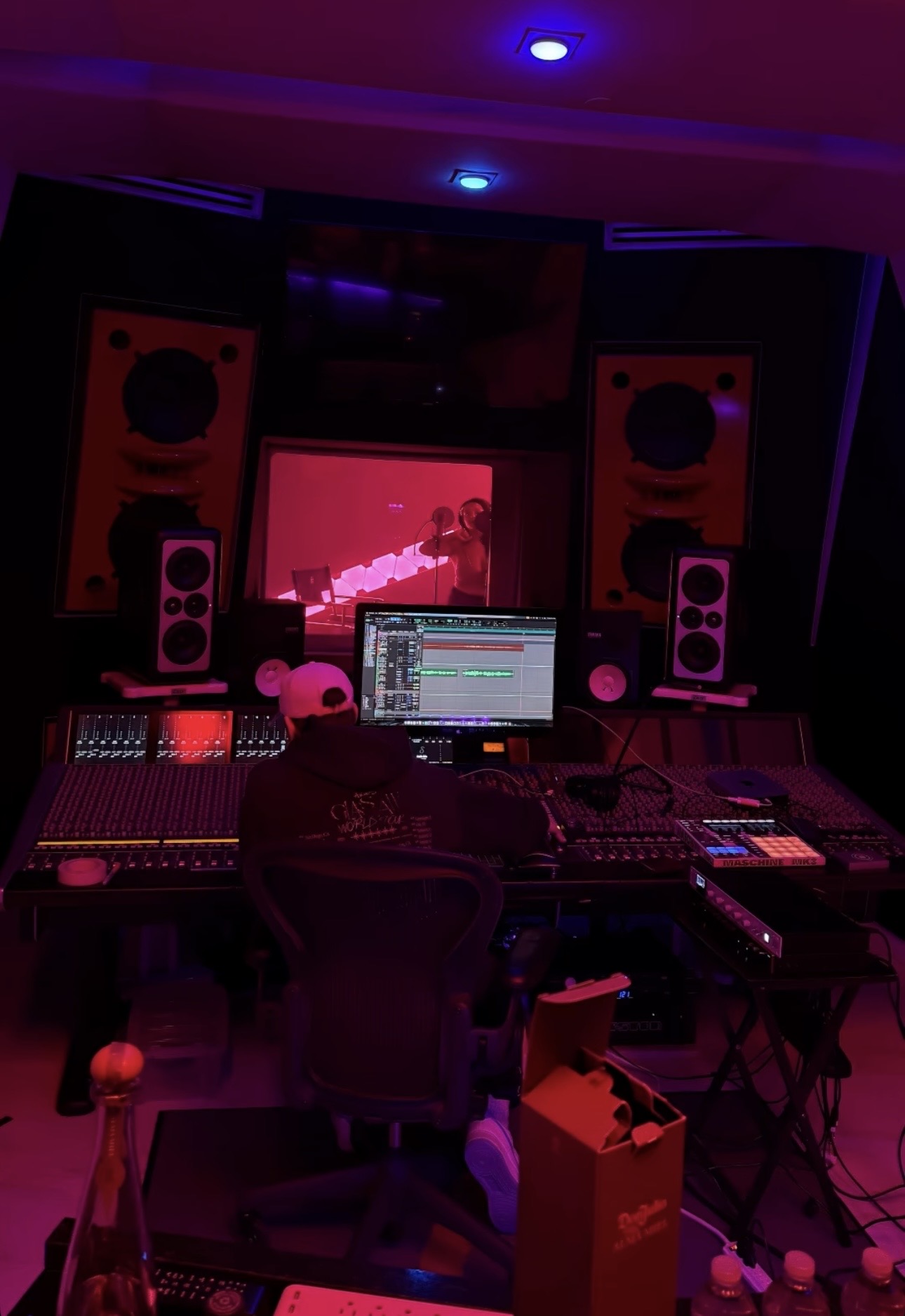 Recording studio with mixing console, computer, speakers, and a person working, illuminated by pink and purple lighting.