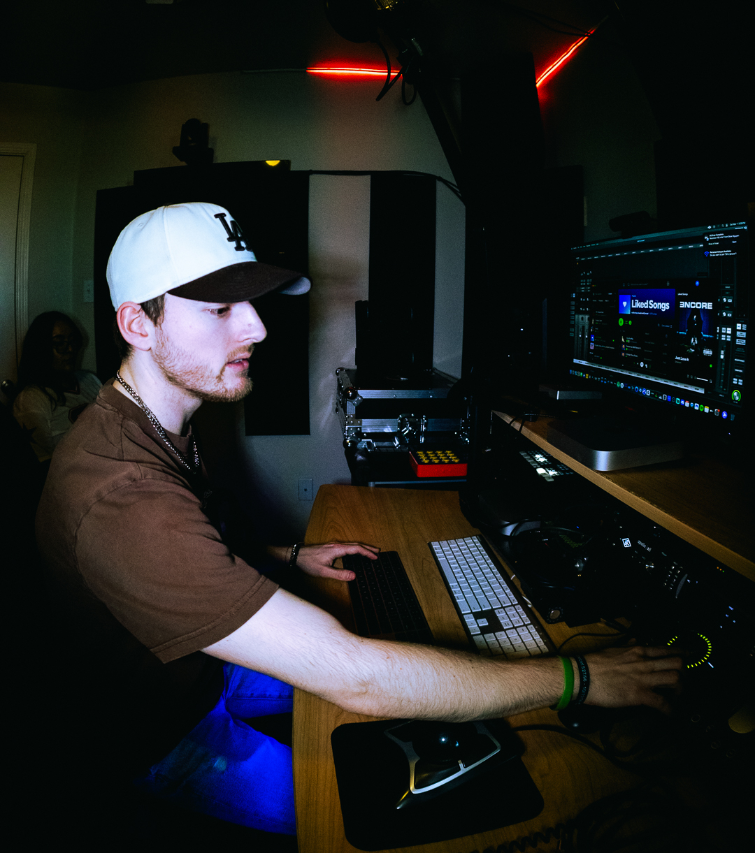 Person wearing a white cap and brown shirt working at a DJ setup with computer and equipment in a dark room.